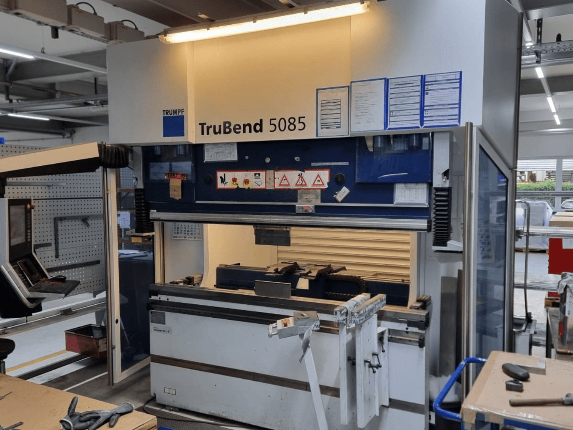 TRUMPF TruBend 5085 liekšanas iekārta, skats no priekšpuses, kurā redzams vadības panelis, darbstacija un drošības brīdinājumi darbnīcā.