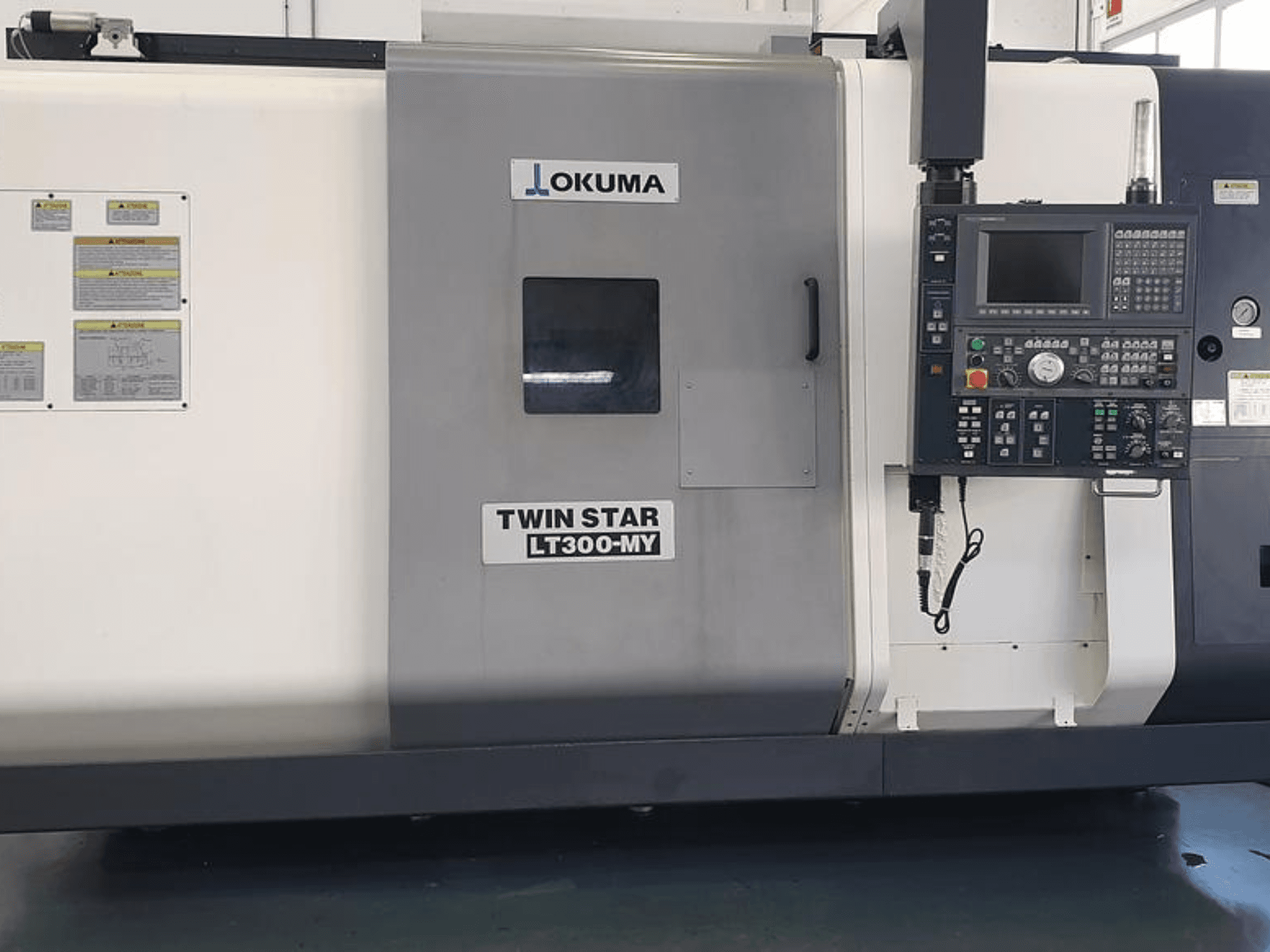 Okuma Twin Star LT300-MY CNC virpa sānu skatā, ar vadības paneli, vadības pogām un drošības marķējumiem.