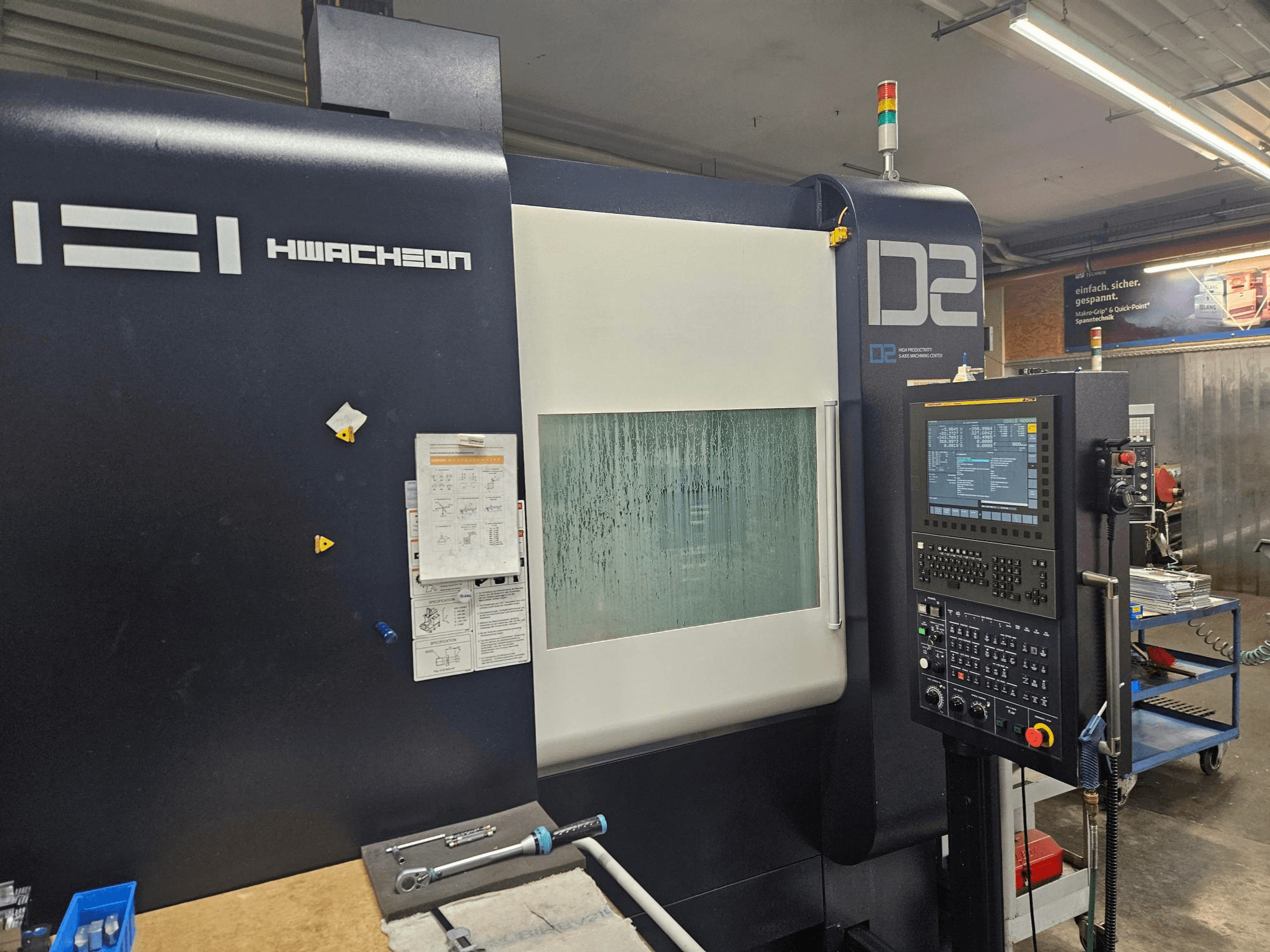 HWACHEON D2-5AX 12k vertikālais apstrādes centrs, skats no priekšpuses, kurā redzama vadības paneļa un aizsūbējies skata logs.