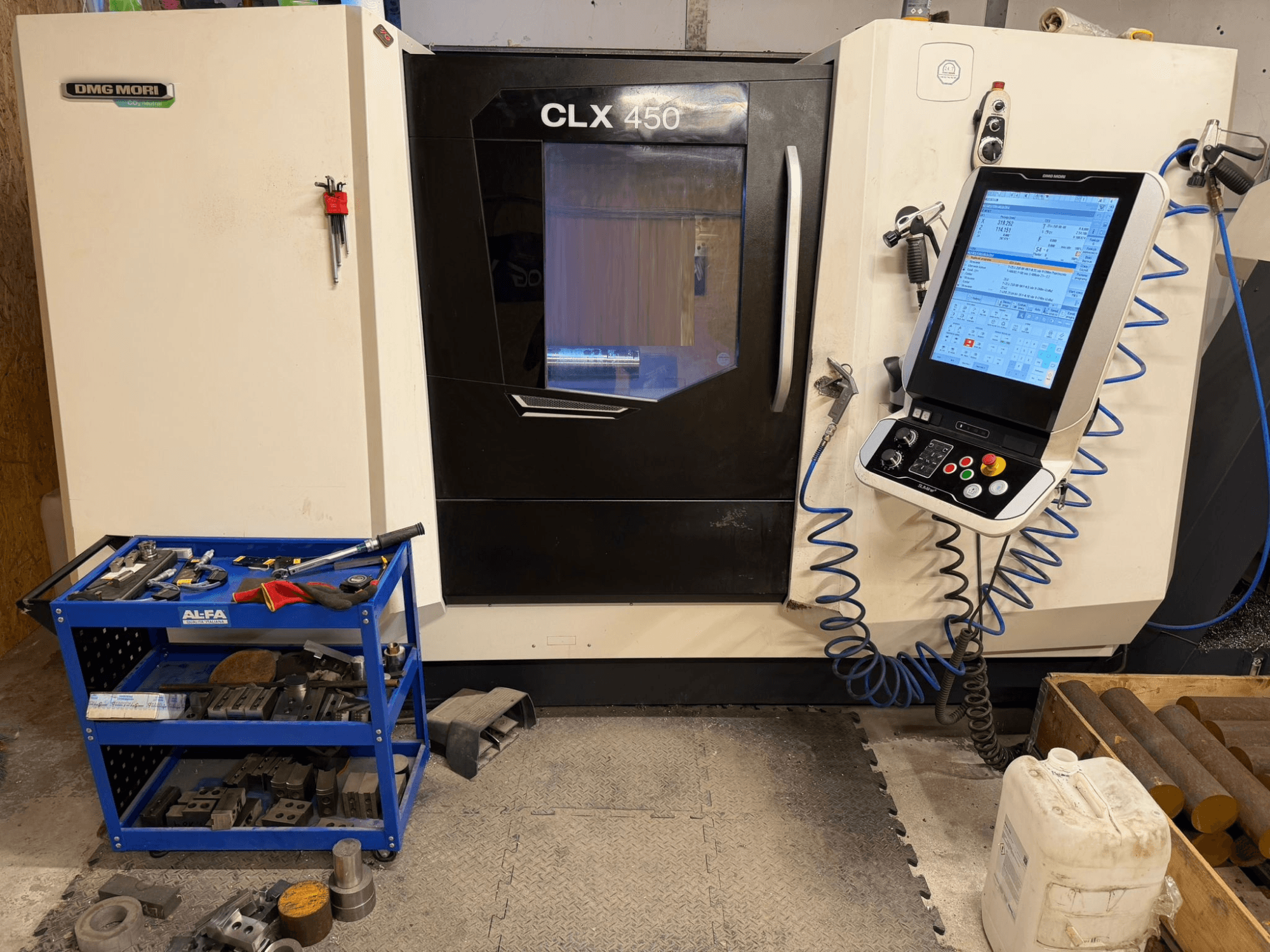 DMG MORI CLX 450 V3 horizontālās virpošanas mašīnas priekšējais skats ar vadības paneli un blakus esošo instrumentu ratiņu ar aprīkojumu.