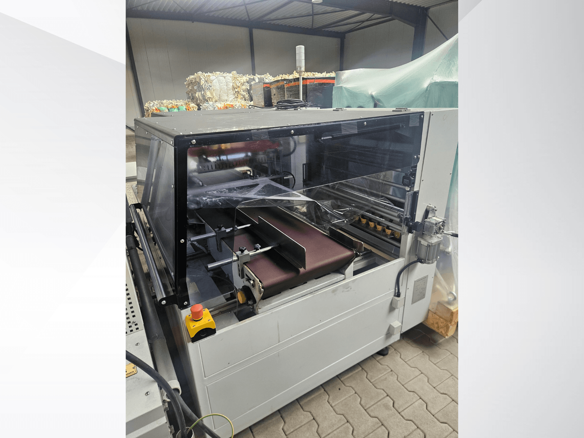 AMTEC Speed Shrink 40 PRO iekārta, skats no priekšpuses, kurā redzams plastmasas apstrādes mehānisms ar caurspīdīgu drošības pārsegu un konveijeru.