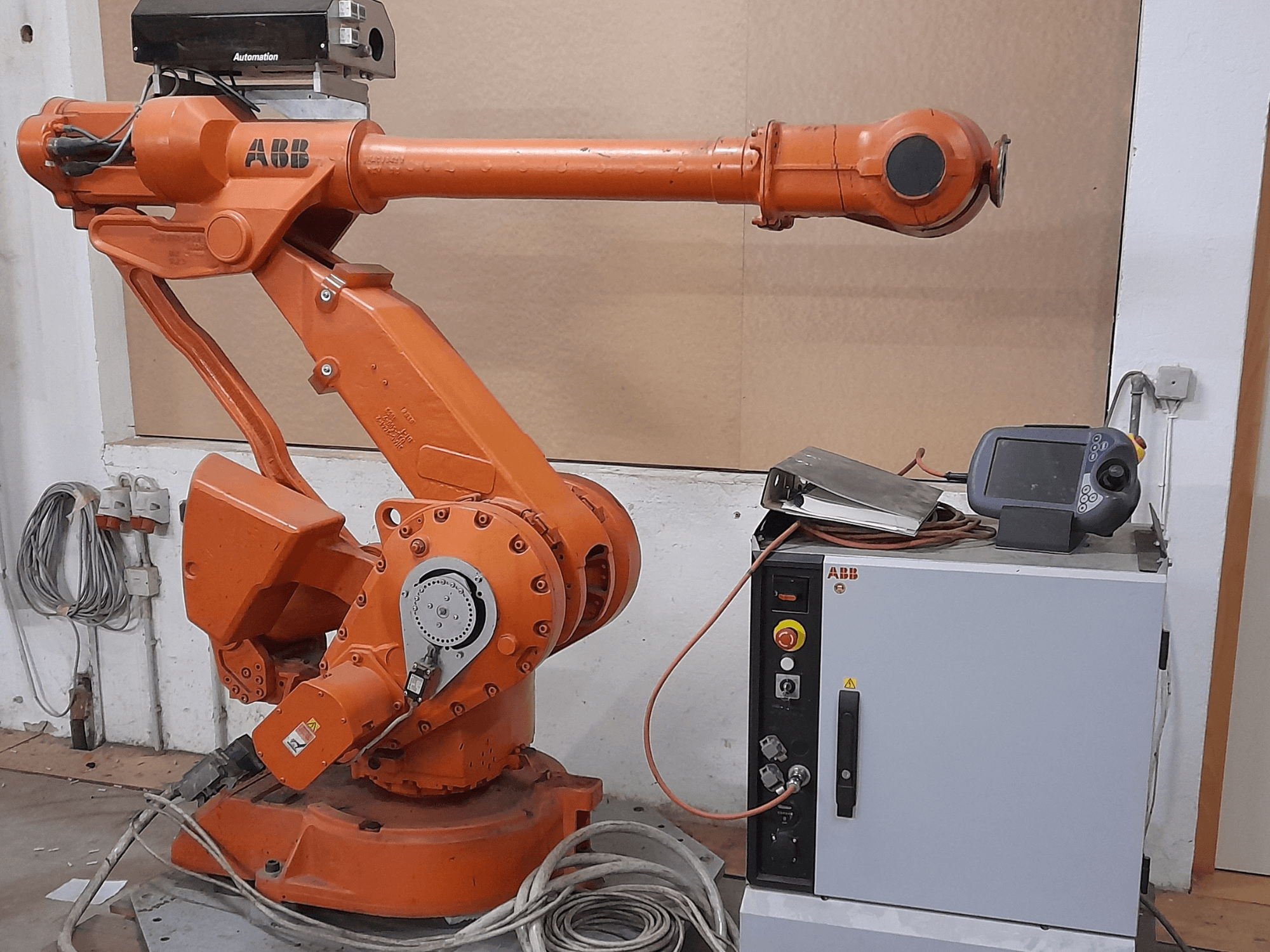 Oranžā ABB robotizētā roka ar vadības paneli, skats no priekšpuses, kurā redzami kabeļi un automatizācijas aprīkojums.