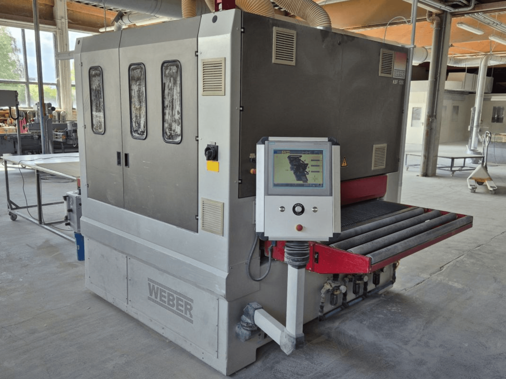 WEBER KSF-3-1350 lentes slīpmašīna, sānu skats, kurā redzamas vadības ierīces, barošanas panelis un slīpēšanas konveijers. Rūpnieciskā vide ar redzamām iekārtām.