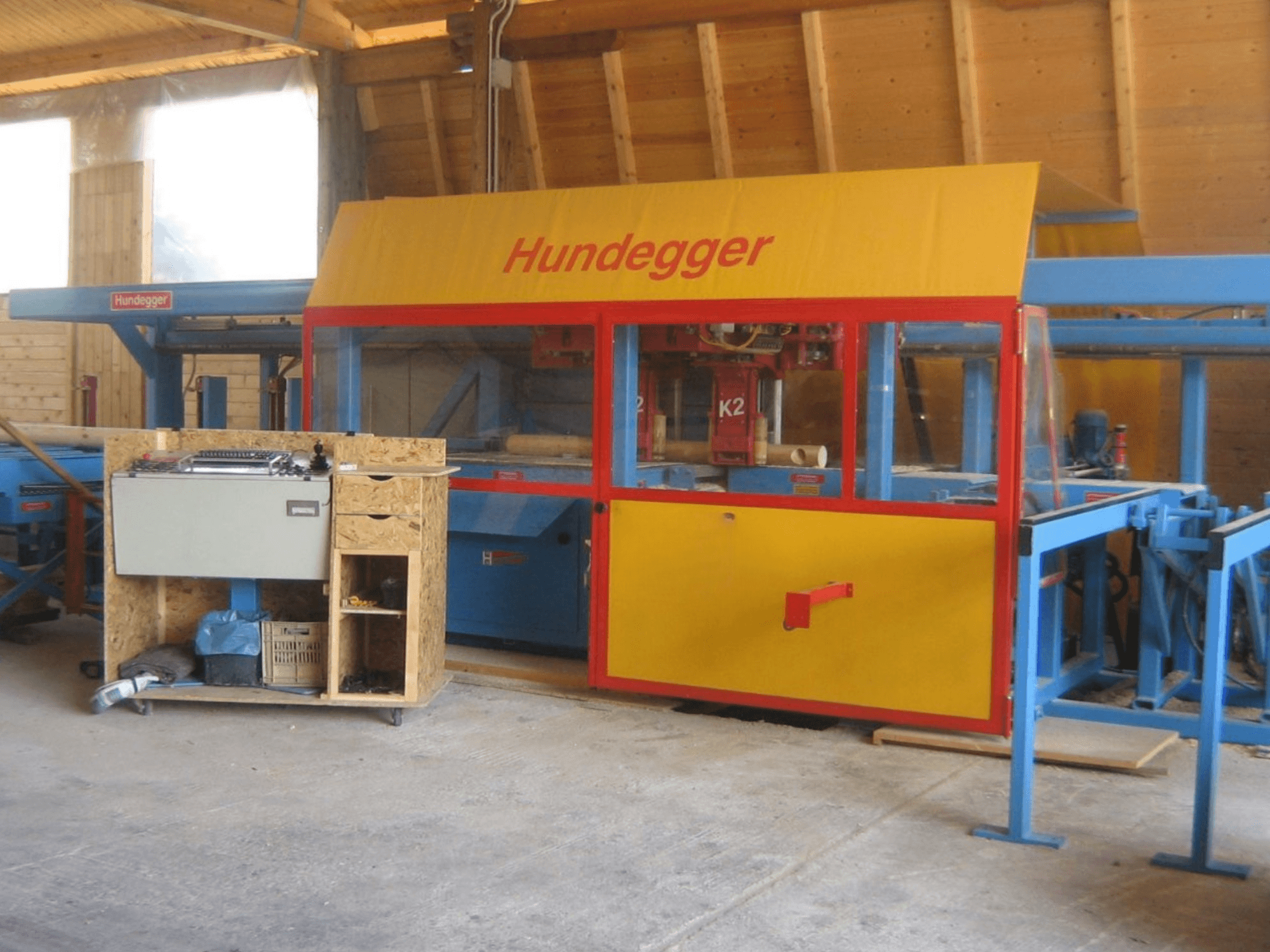 CNC kokapstrādes centrs HUNDEGGER K2 4 assi darbnīcā, priekšējais skats ar dzelteno un zilo dizainu un darba galdu.
