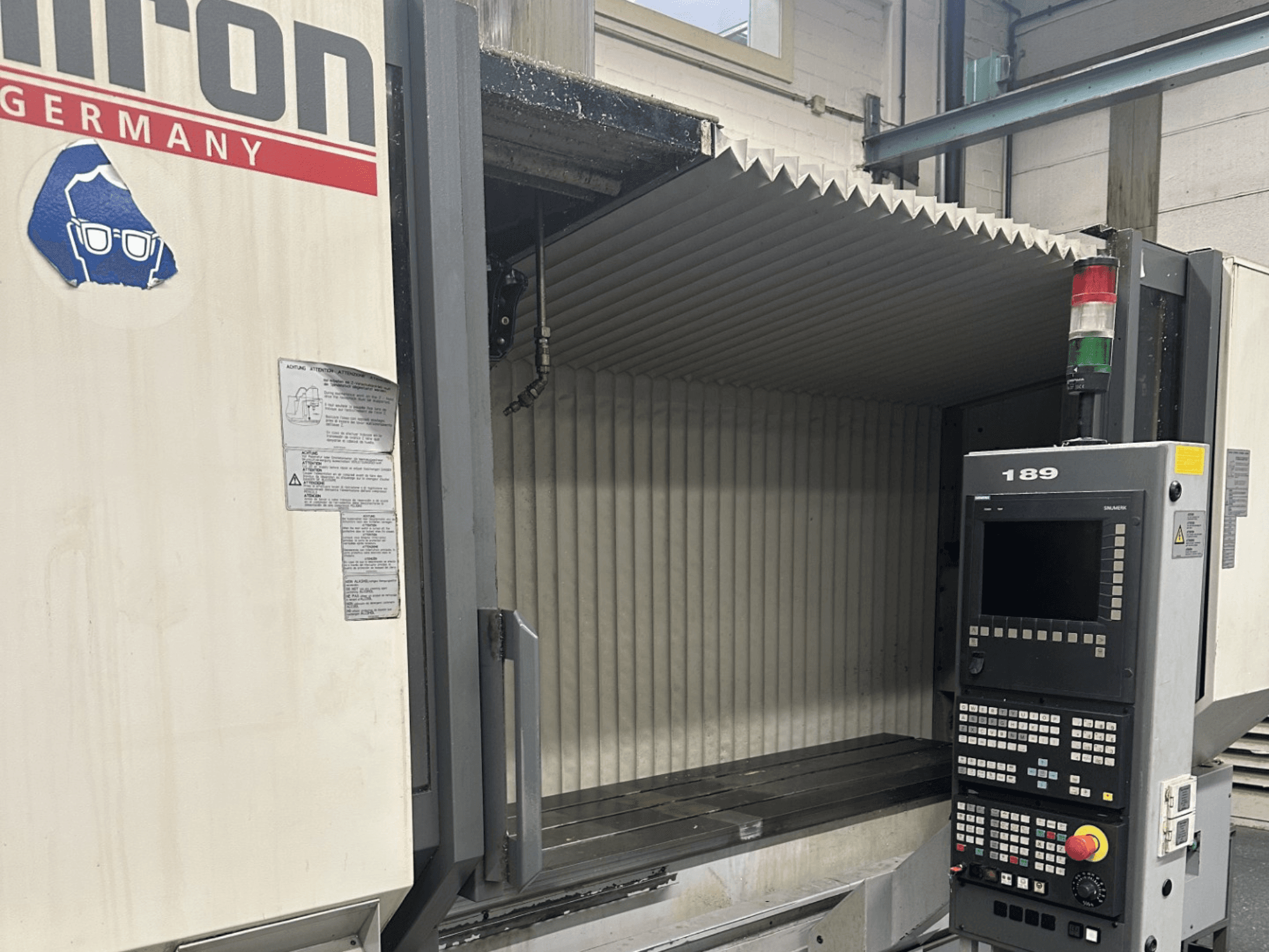 Alt teksts: Tālrunis: Hermle CNC mašīnas priekšējais skats ar vadības paneli, uz kura redzamas pogas un displejs, ar atvērtu darba zonu.