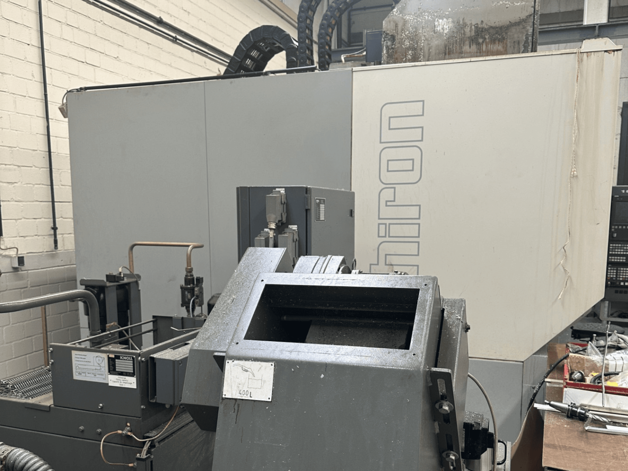 ZĪMOLS: Chiron; Priekšējais skats uz rūpniecisko CNC mašīnu ar pelēku ārpusi, redzams vadības panelis un mašīnu sastāvdaļas fokusā.