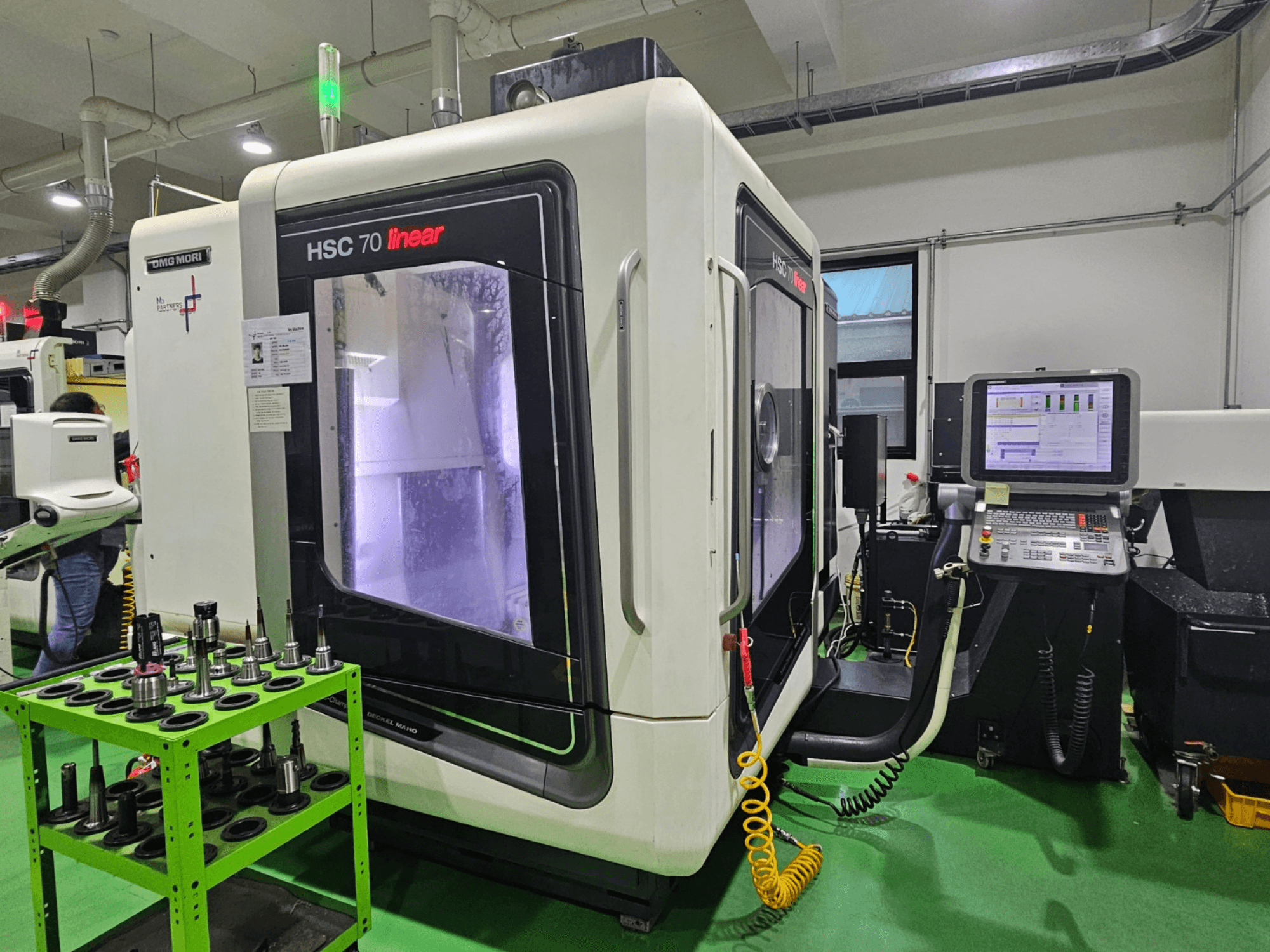 DMG MORI HSC 70 lineārais universālais apstrādes centrs, skats no priekšpuses, ar instrumentu turētājiem uz zaļā galda un redzamu vadības paneli.