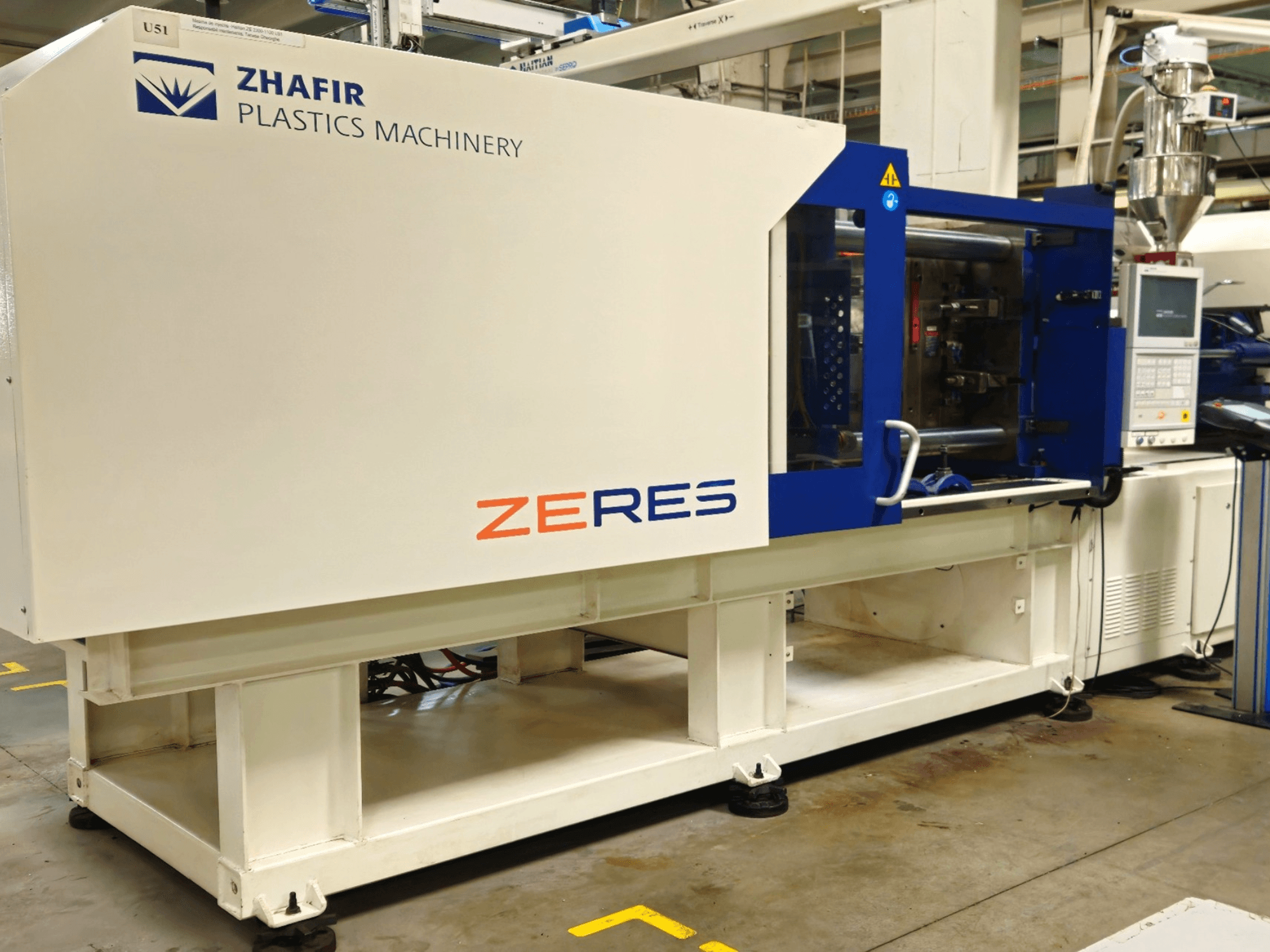 Zhafir Zeres plastmasas iesmidzināšanas iekārta, skats no sāniem, kurā redzams vadības panelis, tvertne un glīts balts/zils dizains.