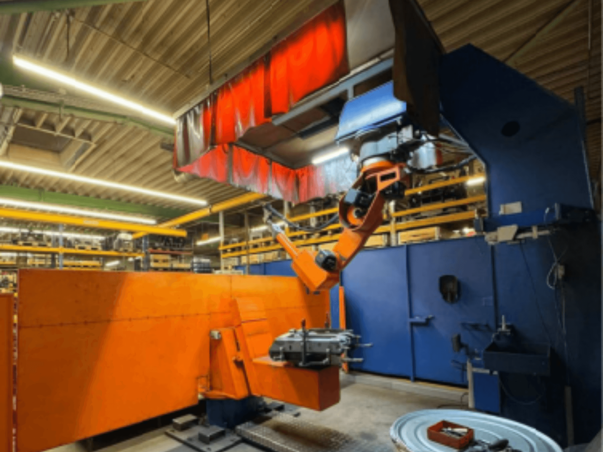 CLOOS Romat 350 e metināšanas robots rūpnieciskā vidē, skats no priekšpuses ar oranžo roku, kas tur apstrādājamo detaļu zem aizkara.