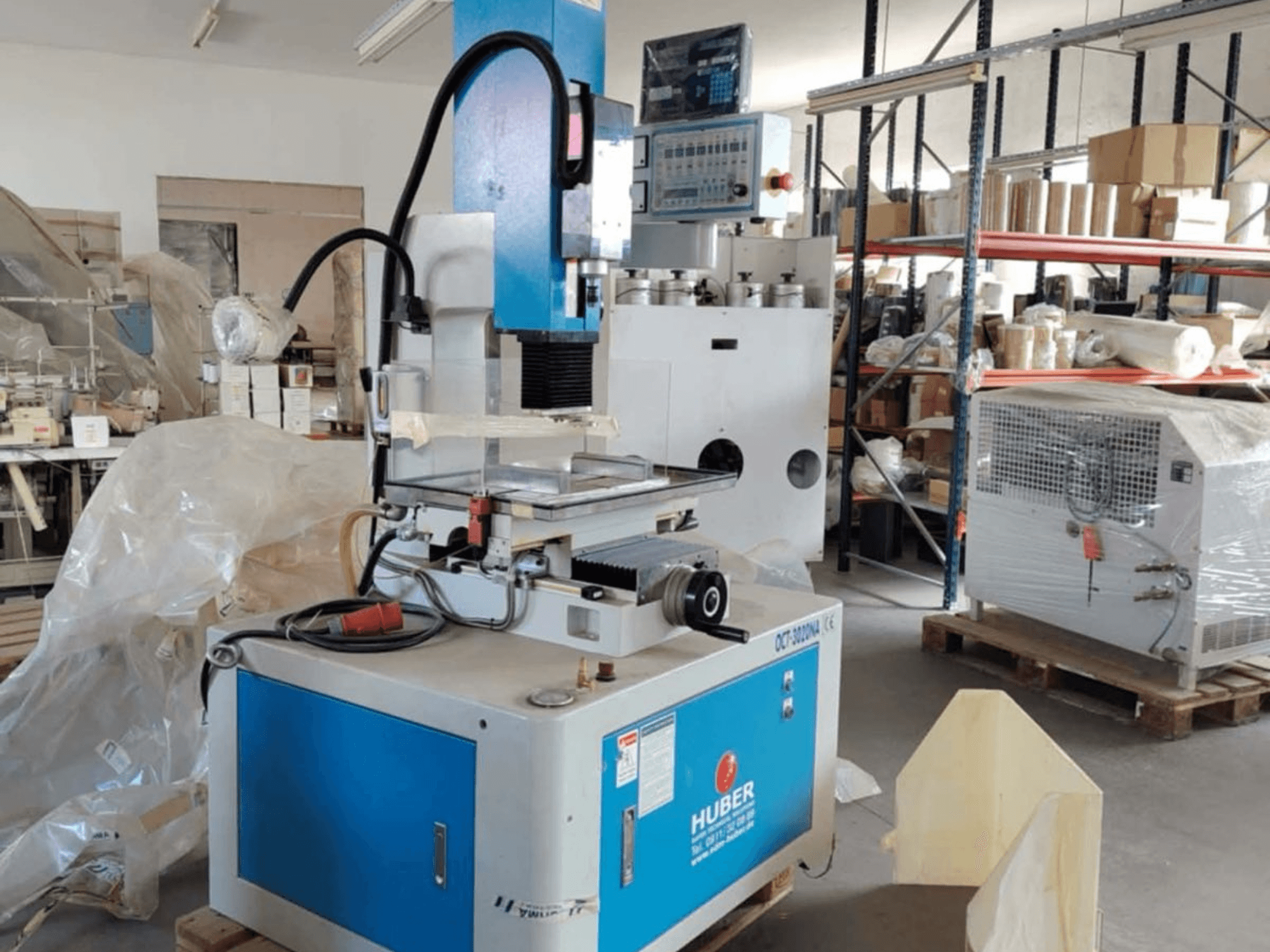 HUBER OCT-3020 NA štancēšanas EDM mašīna darbnīcā, priekšējais skats, kurā redzams zils un pelēks korpuss, digitālās vadības ierīces un darba zona.
