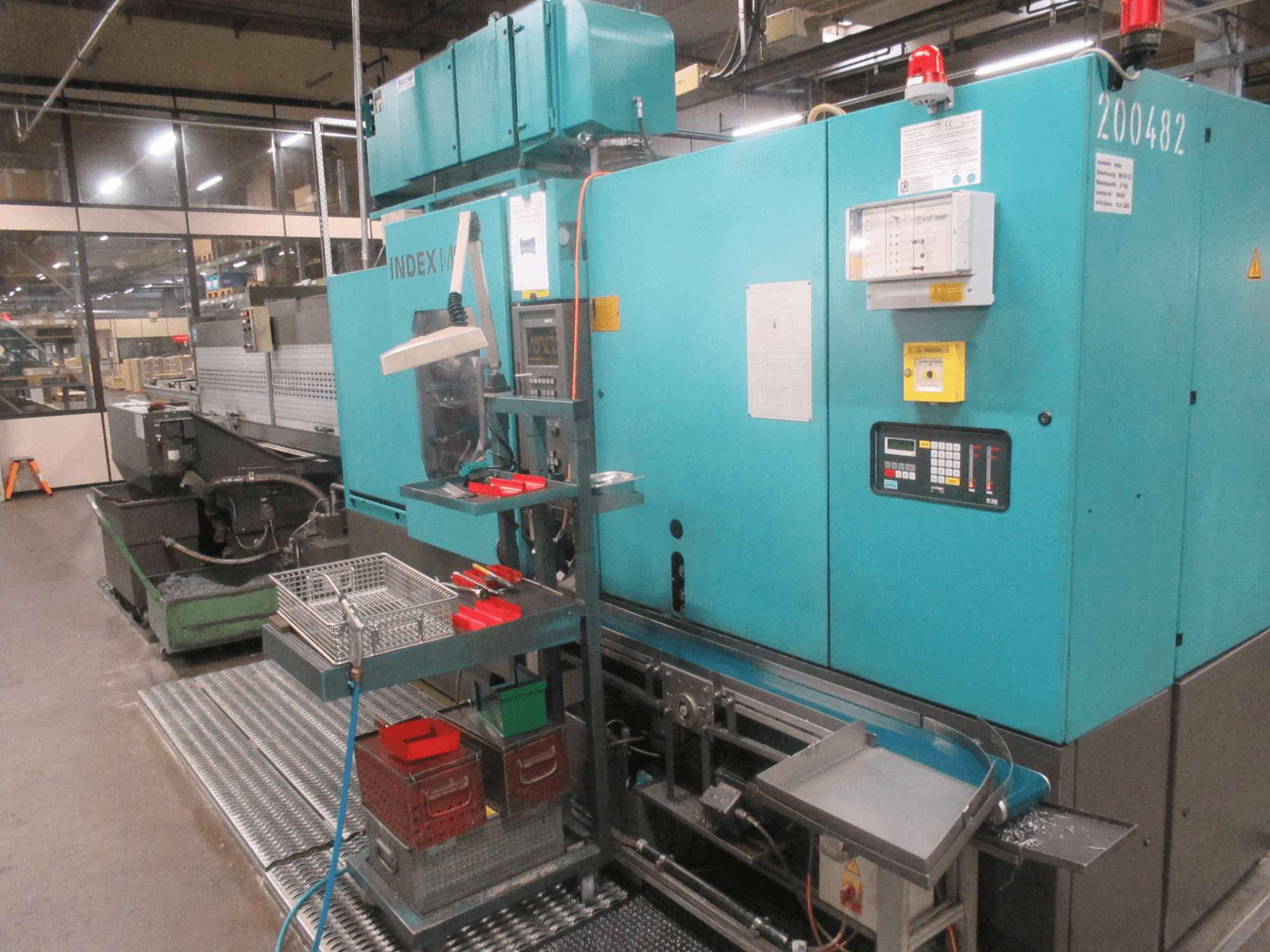 Tirkīza krāsas INDEX mašīna rūpnīcā, skats no sāniem; vadības panelis, instrumentu noliktava un konveijers. Redzamas tvertnes, šļūtenes un signalizācija.