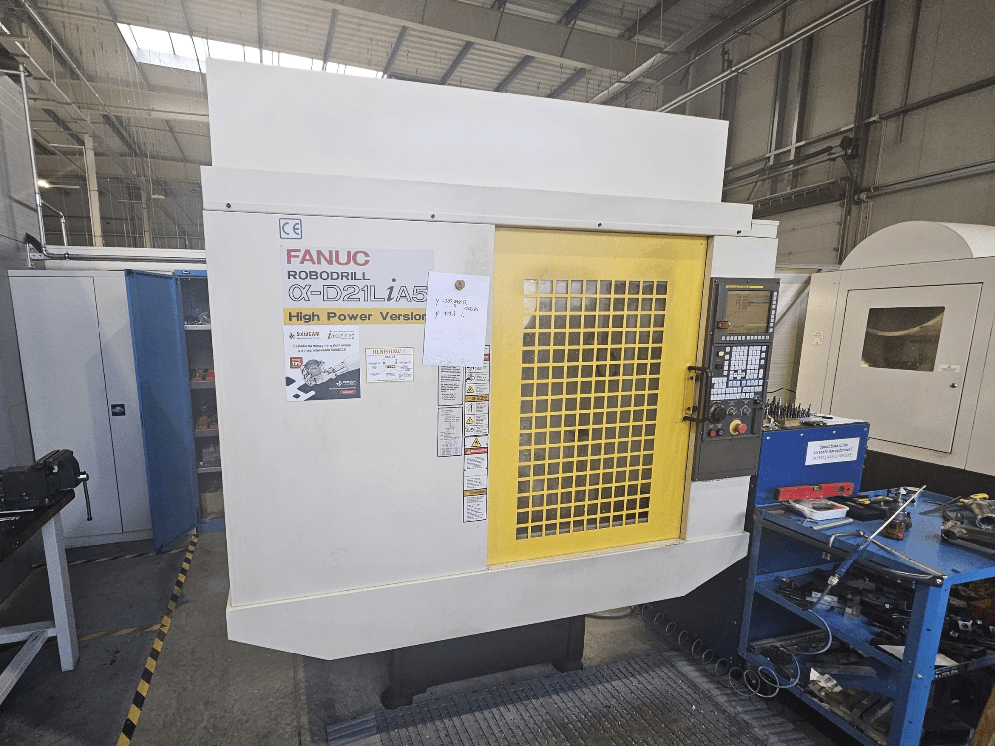 FANUC Robodrill α-D21LiA5 priekšējais skats ar dzeltenām drošības durvīm, vadības paneli un darba vietas instrumentiem tuvumā.