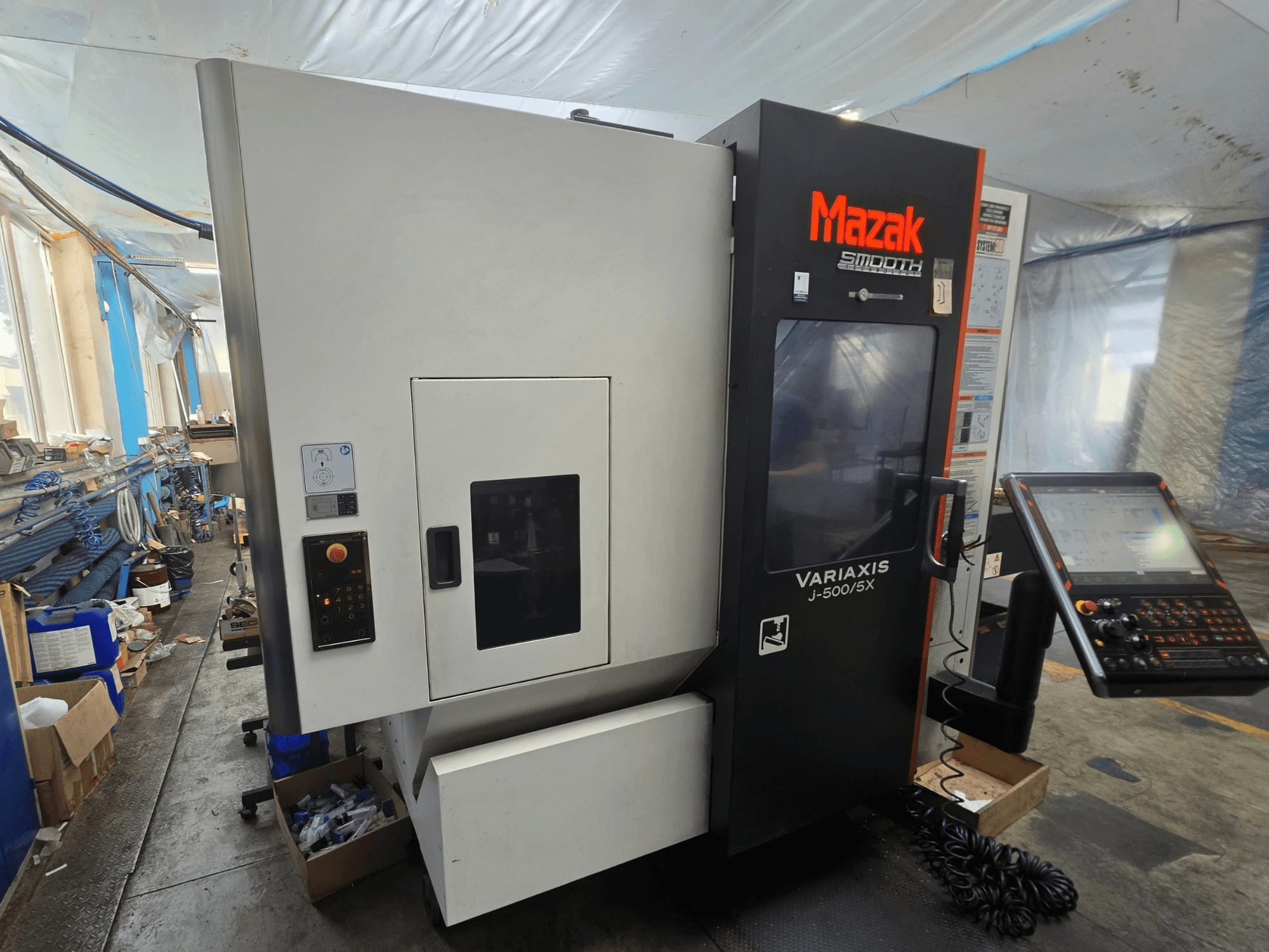 Vertikālais apstrādes centrs „Mazak VARIAXIS J-500/5X“ — sānskats, kurā redzamas vadības ierīces un displejs, uzstādīts darbnīcas vidē.