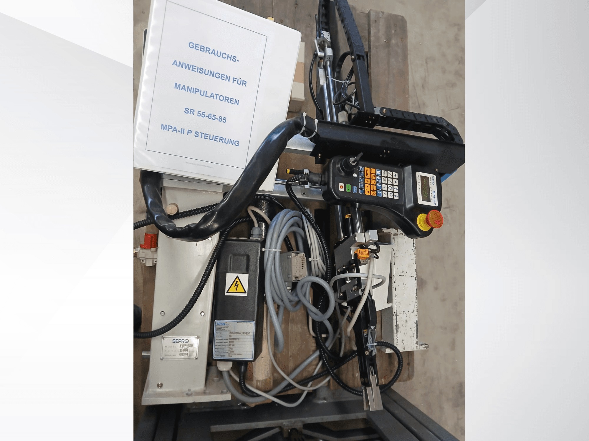 Sepro PIP-55 portālceltnes robota vadības panelis un komponenti, skatoties no sāniem, ar redzamu lietotāja rokasgrāmatu.