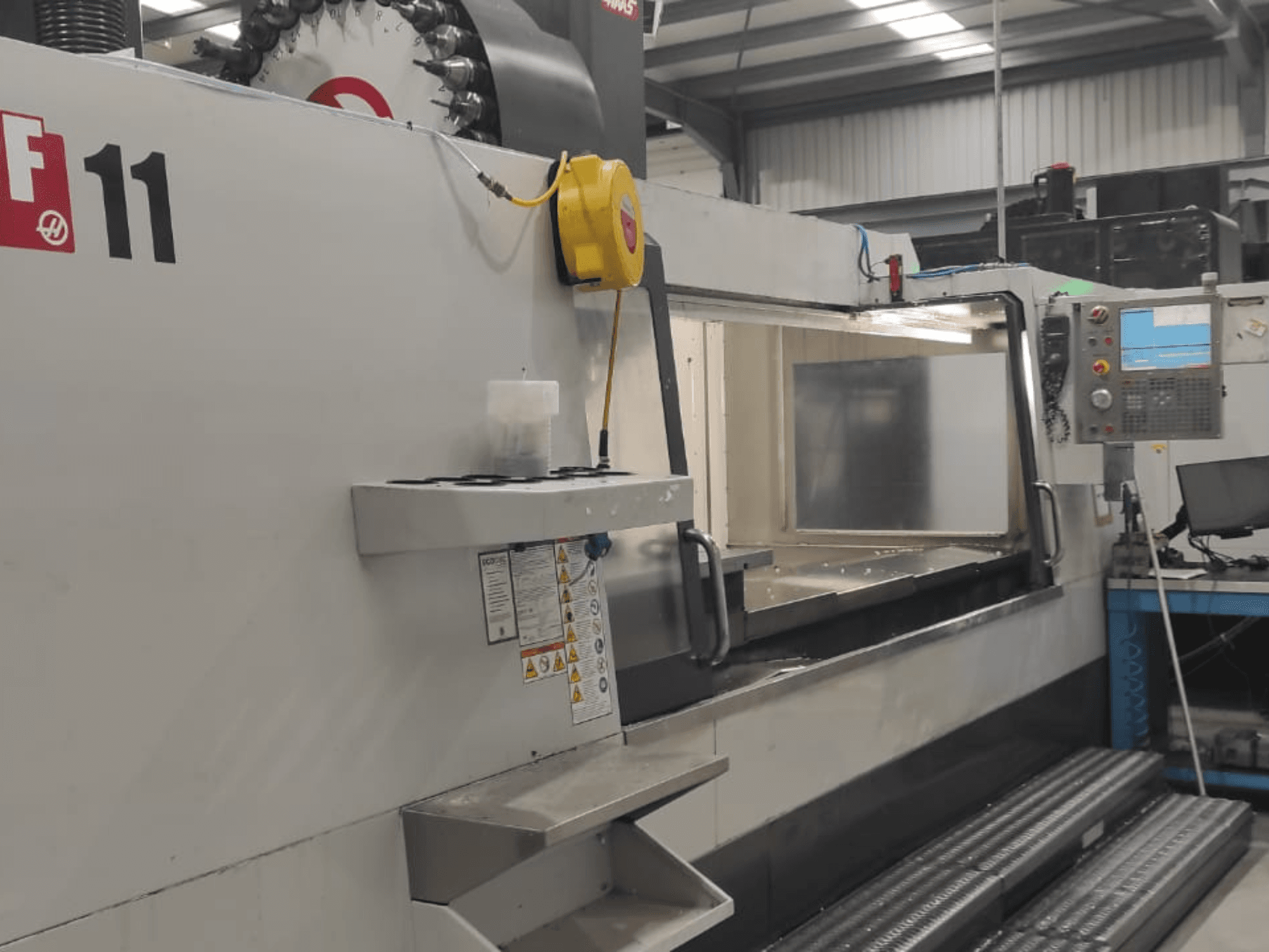 Haas mašīna, modelis VF-11, parādīta no priekšpuses, redzams vadības panelis, liela darba zona un drošības marķējums.