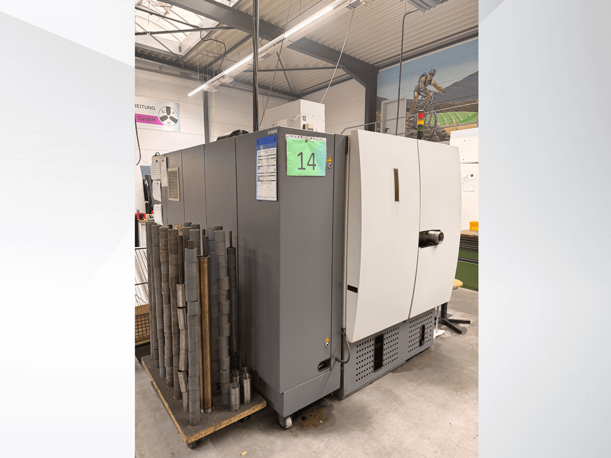 Siemens pelēkā rūpnieciskā mašīna, skats no sāniem, ar cilindrisko metāla gabalu krājumu blakus un redzamām iekārtu etiķetēm.