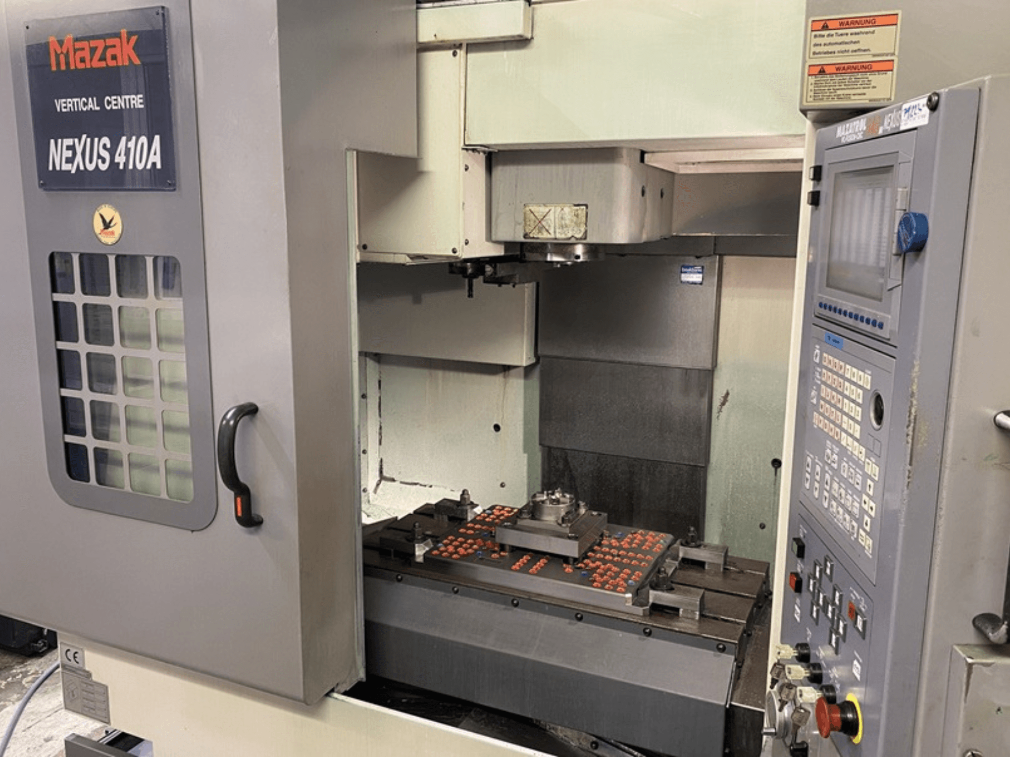 Mazak VCN410 vertikālās apstrādes centrs, priekšējais skats, kurā redzams vadības panelis un mašīnas iekšpuse ar uzstādītu apstrādājamo detaļu.