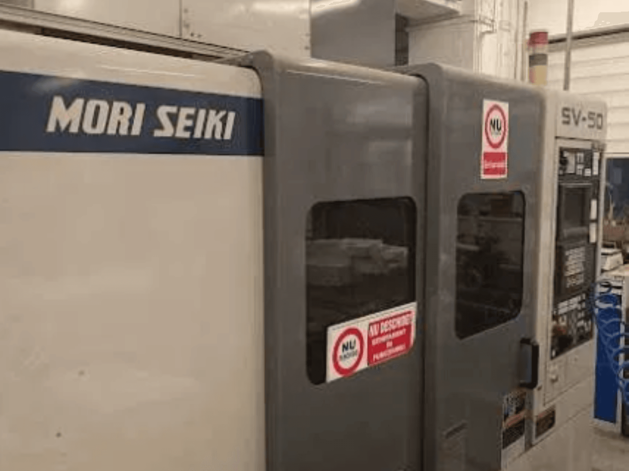 MORI SEIKI SV-50 CNC mašīna, skats no sāniem; vadības panelis un drošības brīdinājuma uzlīmes.
