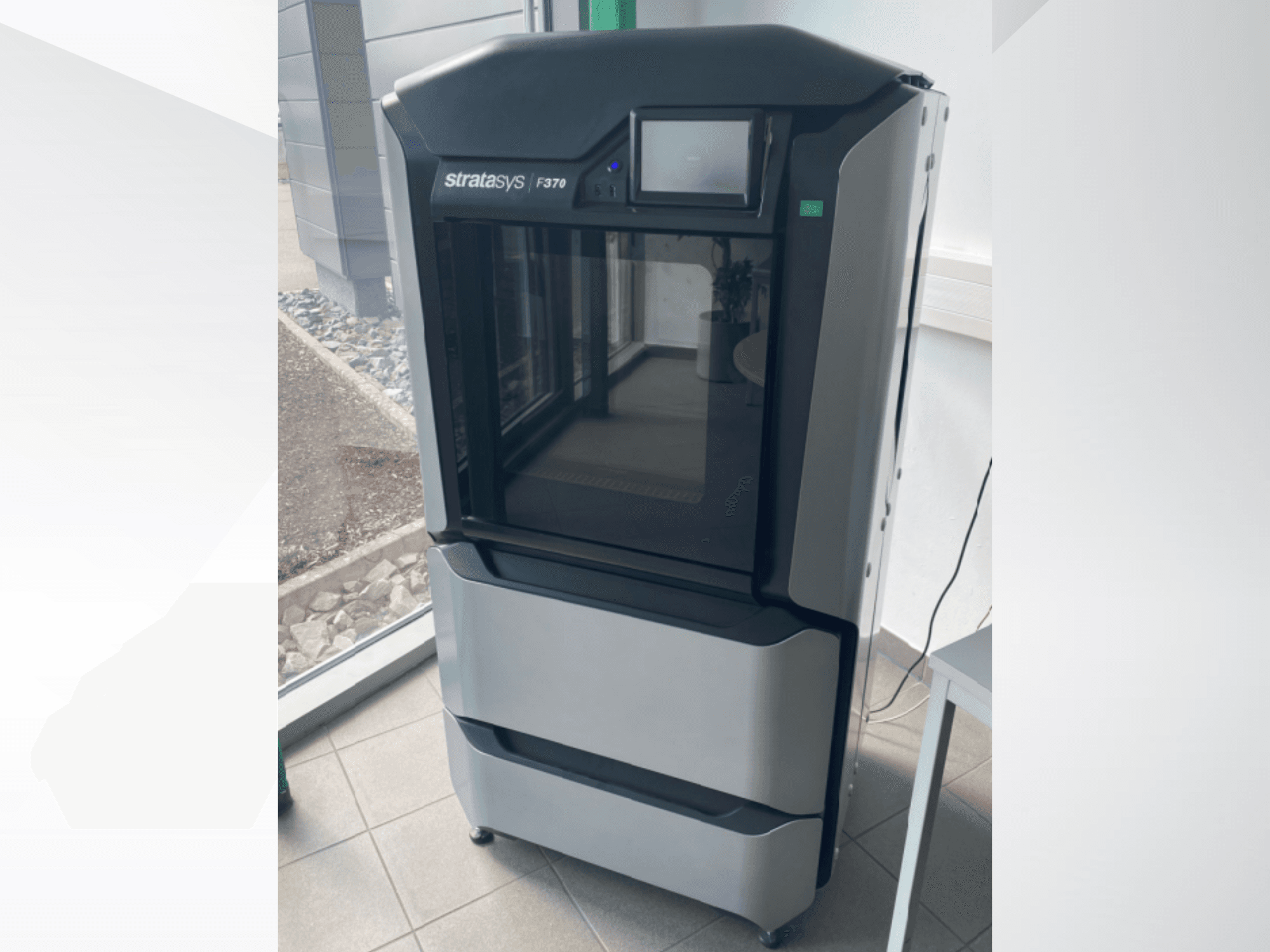 Stratasys F370 plastmasas 3D printeris, skats no priekšpuses, kurā redzams glīts melns un pelēks dizains ar skārienjūtīgo ekrānu.
