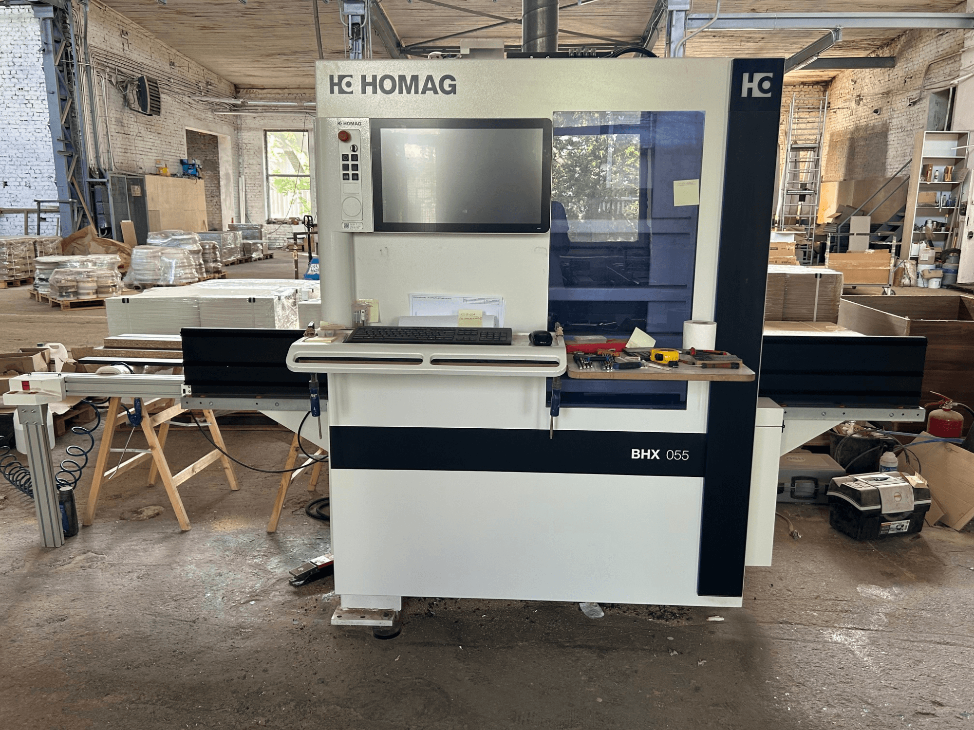 HOMAG WEEKE OPTIMAT BHX 055 CNC apstrādes centrs, skats no priekšpuses, kurā redzams vadības panelis un darba laukums ar instrumentiem.