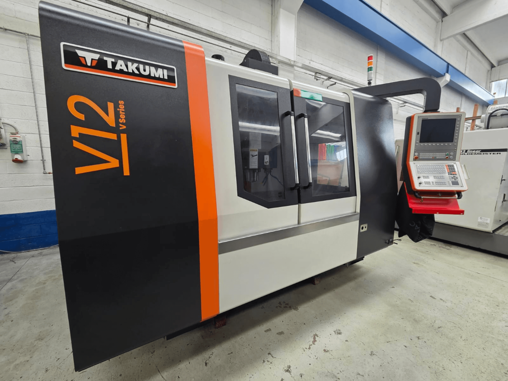 Takumi V12 V sērijas CNC frēzmašīna, skats no priekšpuses, ar vadības paneli un lieliem stikla logiem.