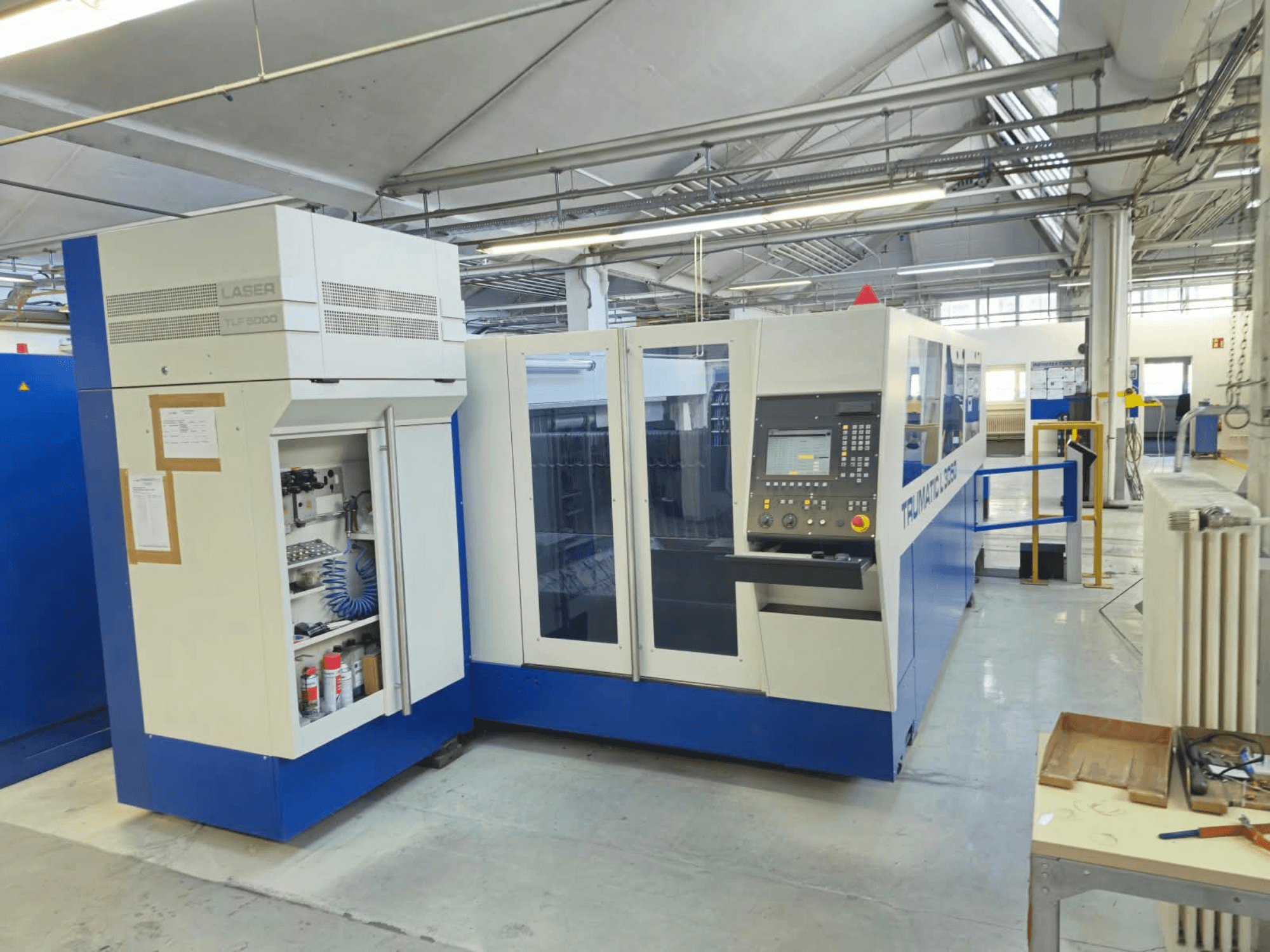 TRUMPF TruLaser L3050 CO₂ lāzergriešanas iekārta, skats no priekšpuses, kurā redzams vadības panelis un apkopes durvis, rūpnīcas konfigurācijā.