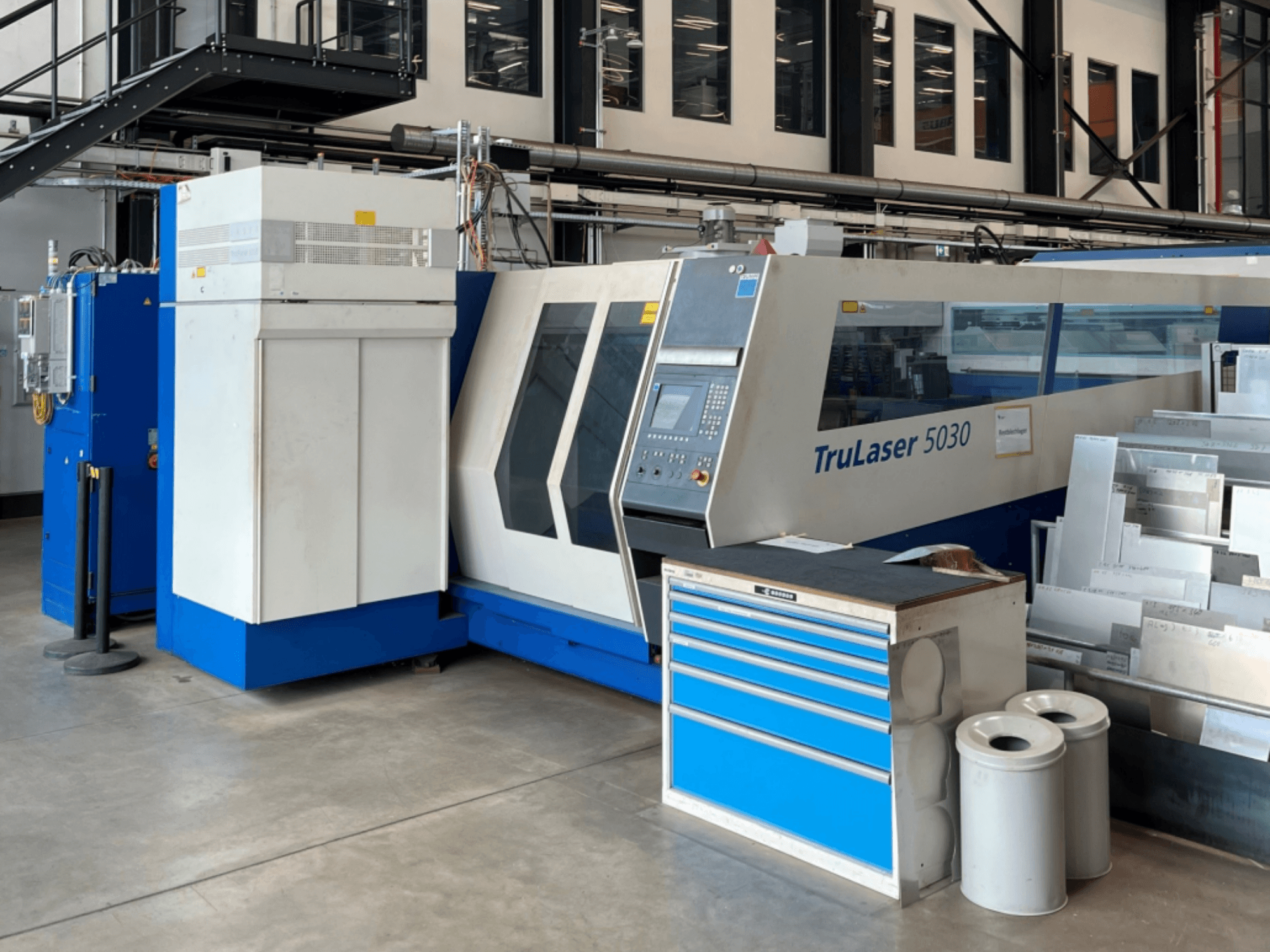 TRUMPF TruLaser 5030 CO₂ lāzergriešanas iekārta, skats no priekšpuses, kurā redzams vadības panelis un blakus esošais darbgalds ar metāla loksnēm.