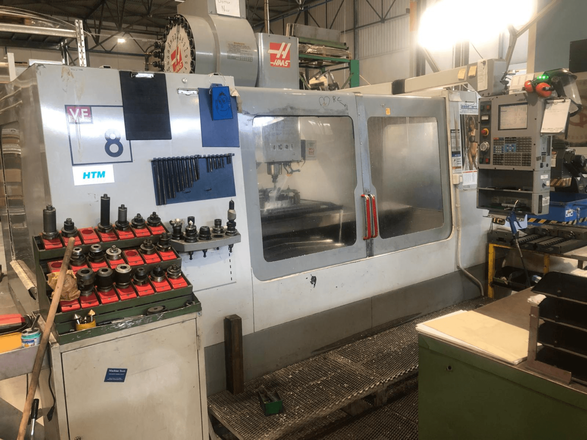 Haas CNC darbnīcā, skats no priekšpuses; redzami instrumenti un vadības ierīces, redzamas modeļa detaļas un sakārtoti instrumentu turētāji.