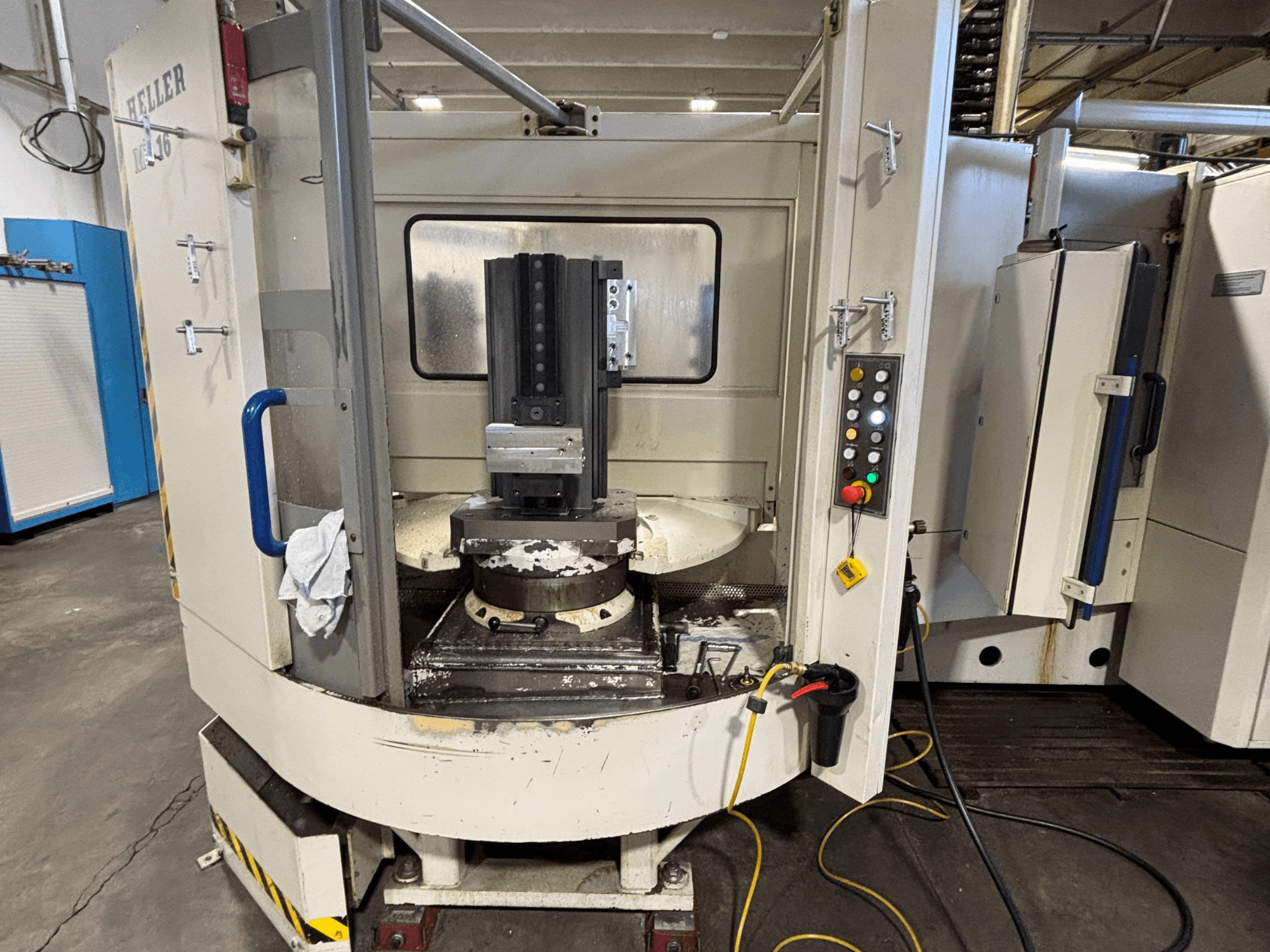 Heller vertikālais apstrādes centrs, vertikālais modelis, priekšējais skats, kurā redzams vārpsta, vadības panelis un darba galds ar piestiprināto aprīkojumu.