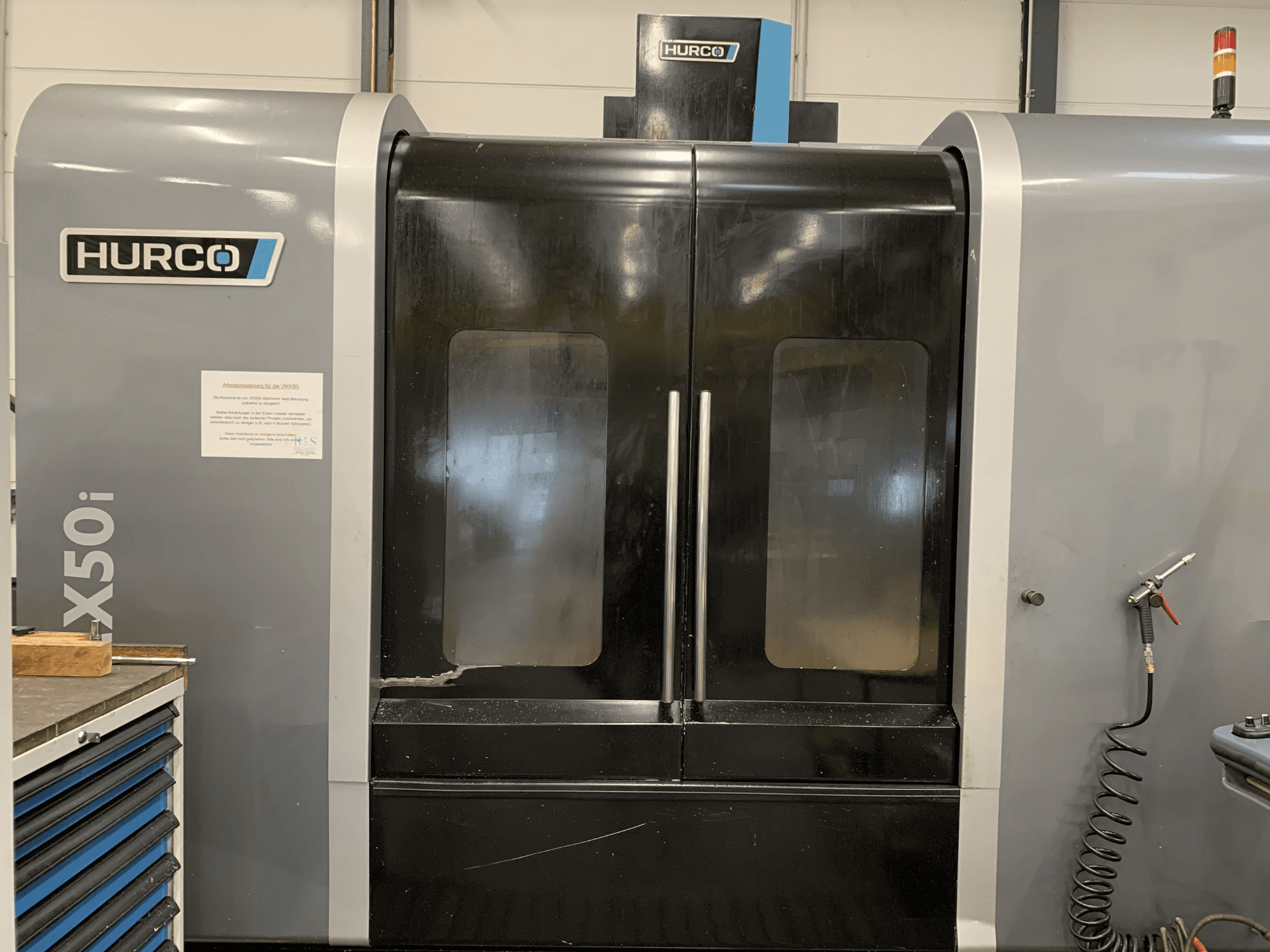 Hurco VX50i CNC mašīnas ar divām durvīm priekšējais skats, tehniskās vadības ierīces un tehniskās apkopes zīme uz sāniem.