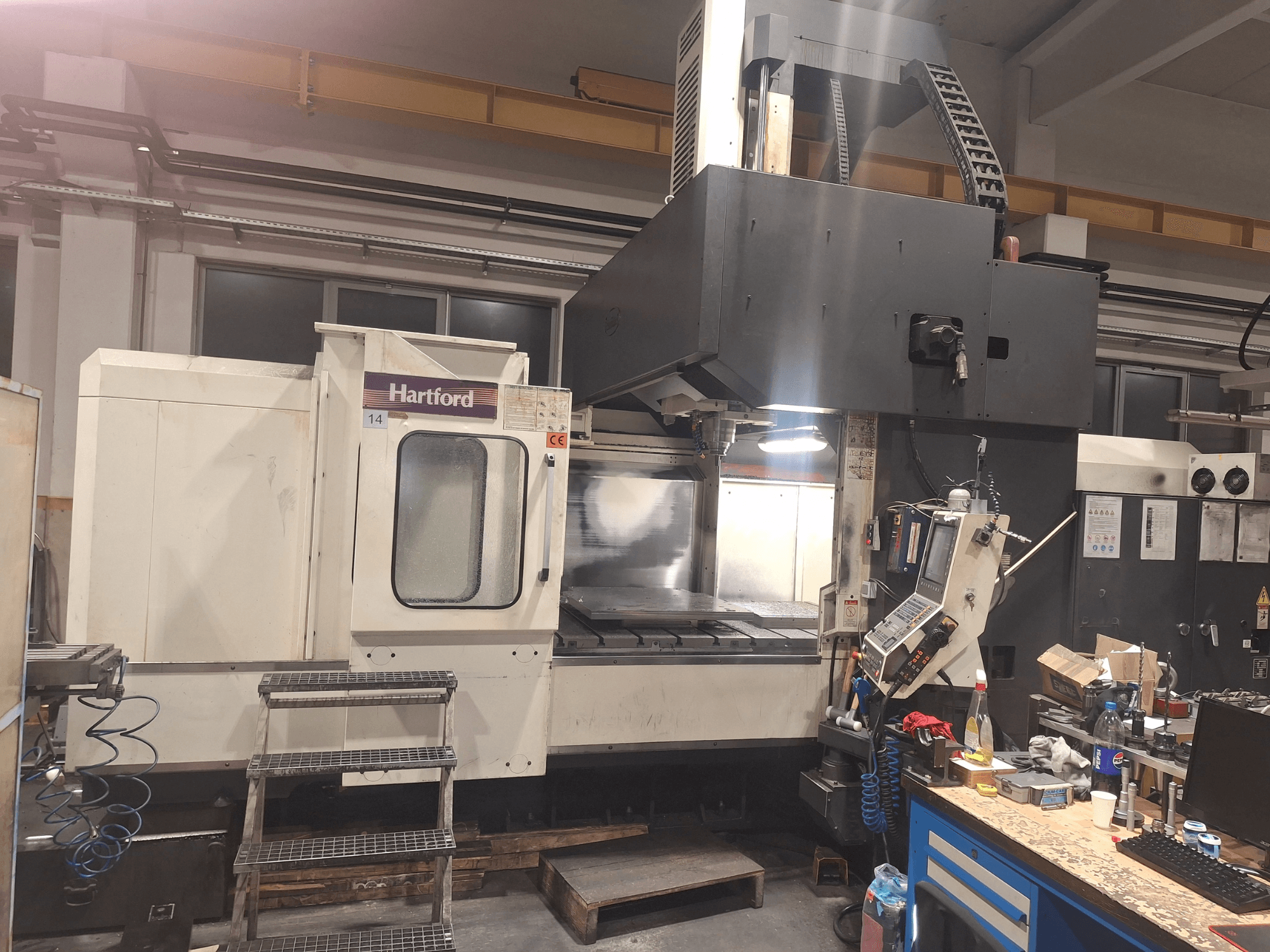 Hartford CNC mašīna, skats no priekšpuses, ar tehniskajām vadības ierīcēm, pakāpieniem, pa kuriem var piekļūt, un darba galda laukumu darbnīcā.
