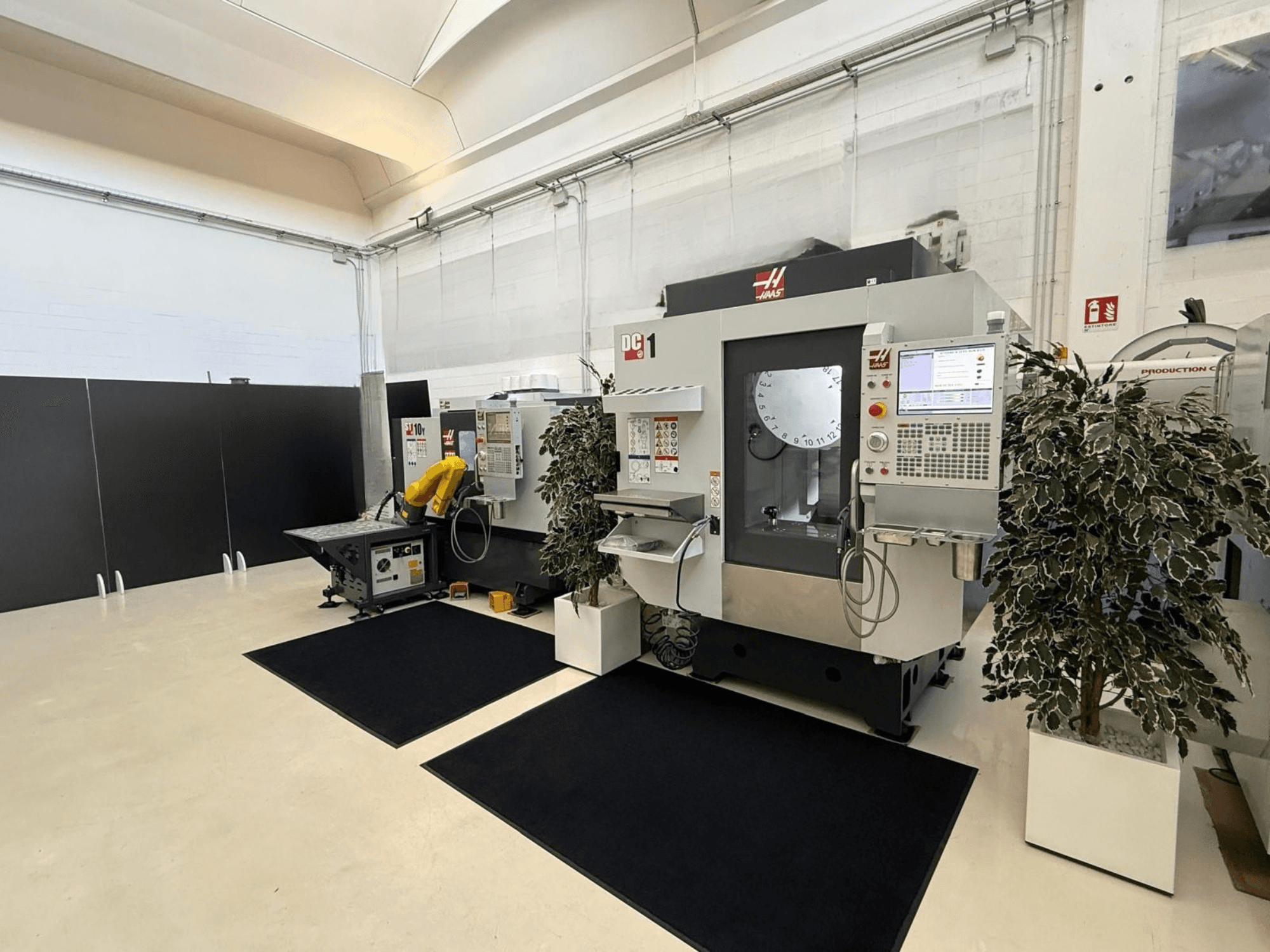 HAAS DC-1 vertikālā apstrādes centra skats no priekšpuses tīrā darba vidē, kurā redzams skārienjūtīgais vadības panelis un apkārt esošās iekārtas.