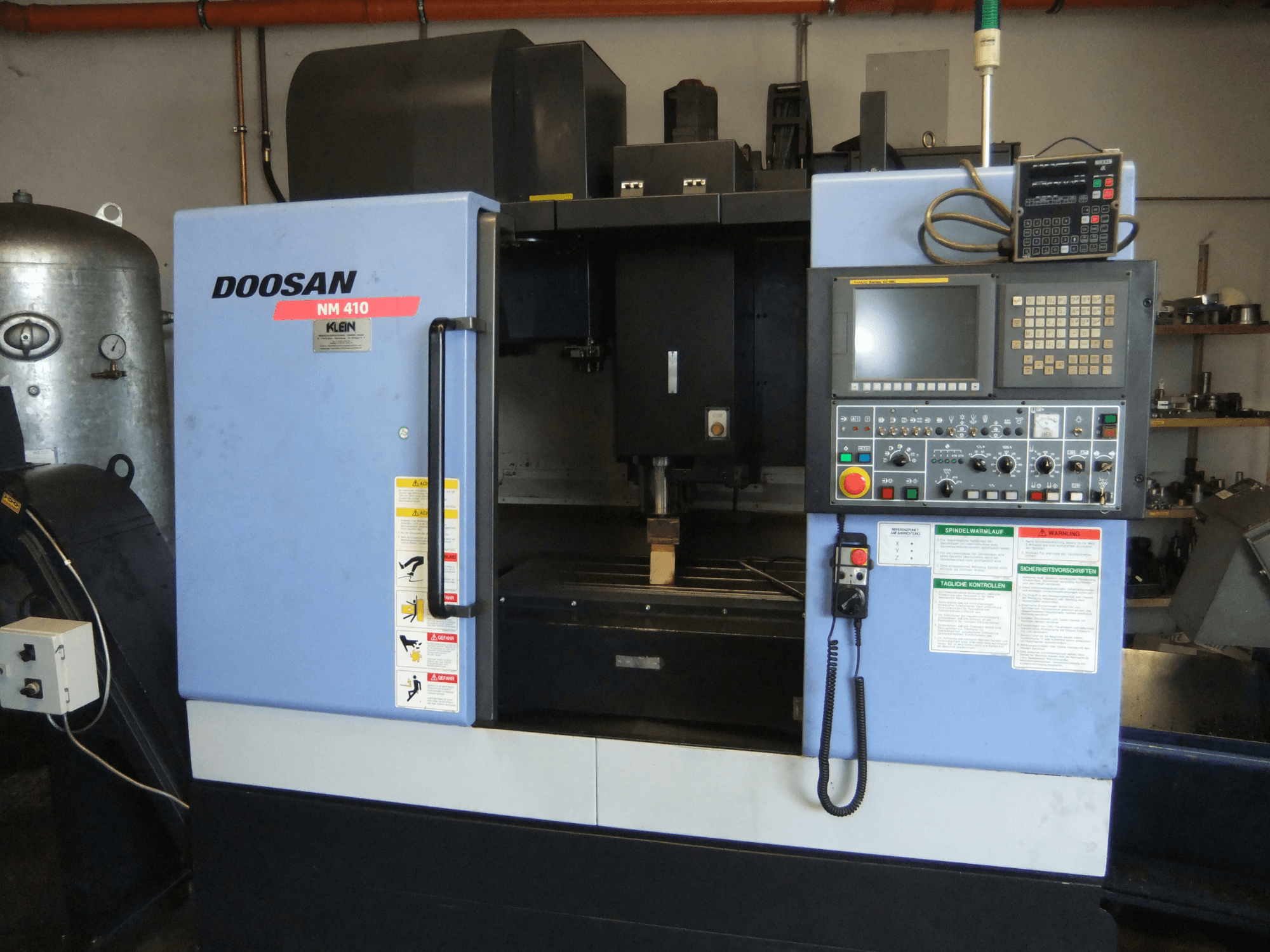 Vertikālais apstrādes centrs DOOSAN NM 410, skats no priekšpuses, kurā redzams vadības panelis, vārpstas zona un drošības uzlīmes.