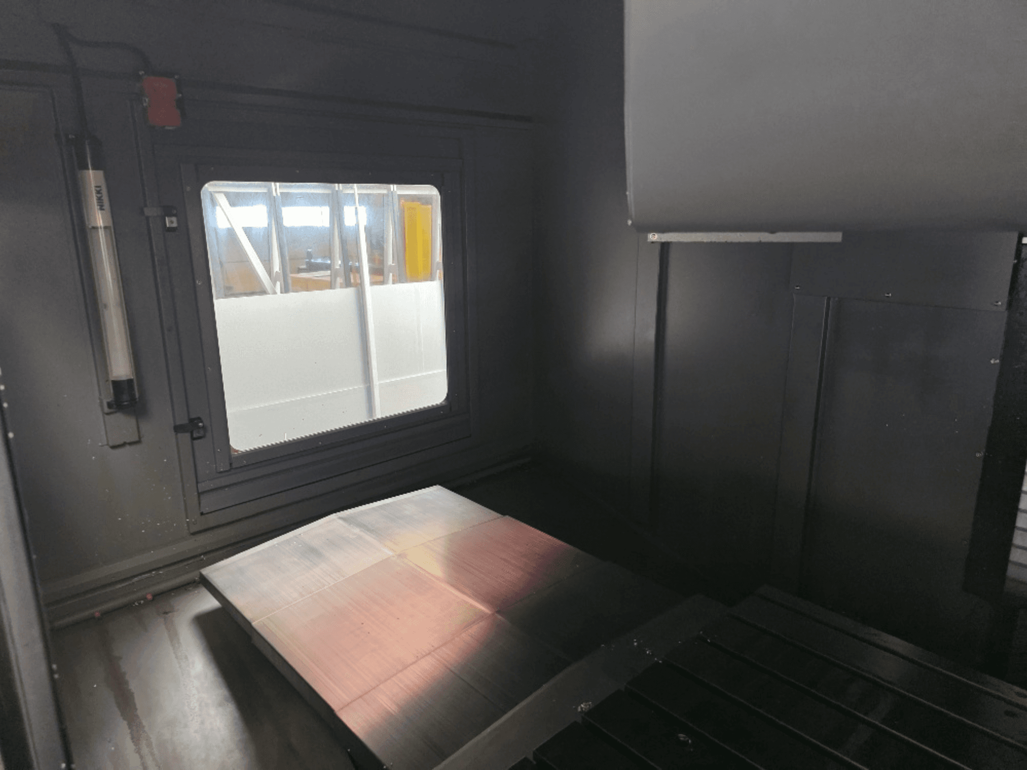 Mašīnas korpusa iekšējais skats ar pulēta metāla plāksni, sānu logu un minimālu apgaismojumu. Nav redzams zīmols.