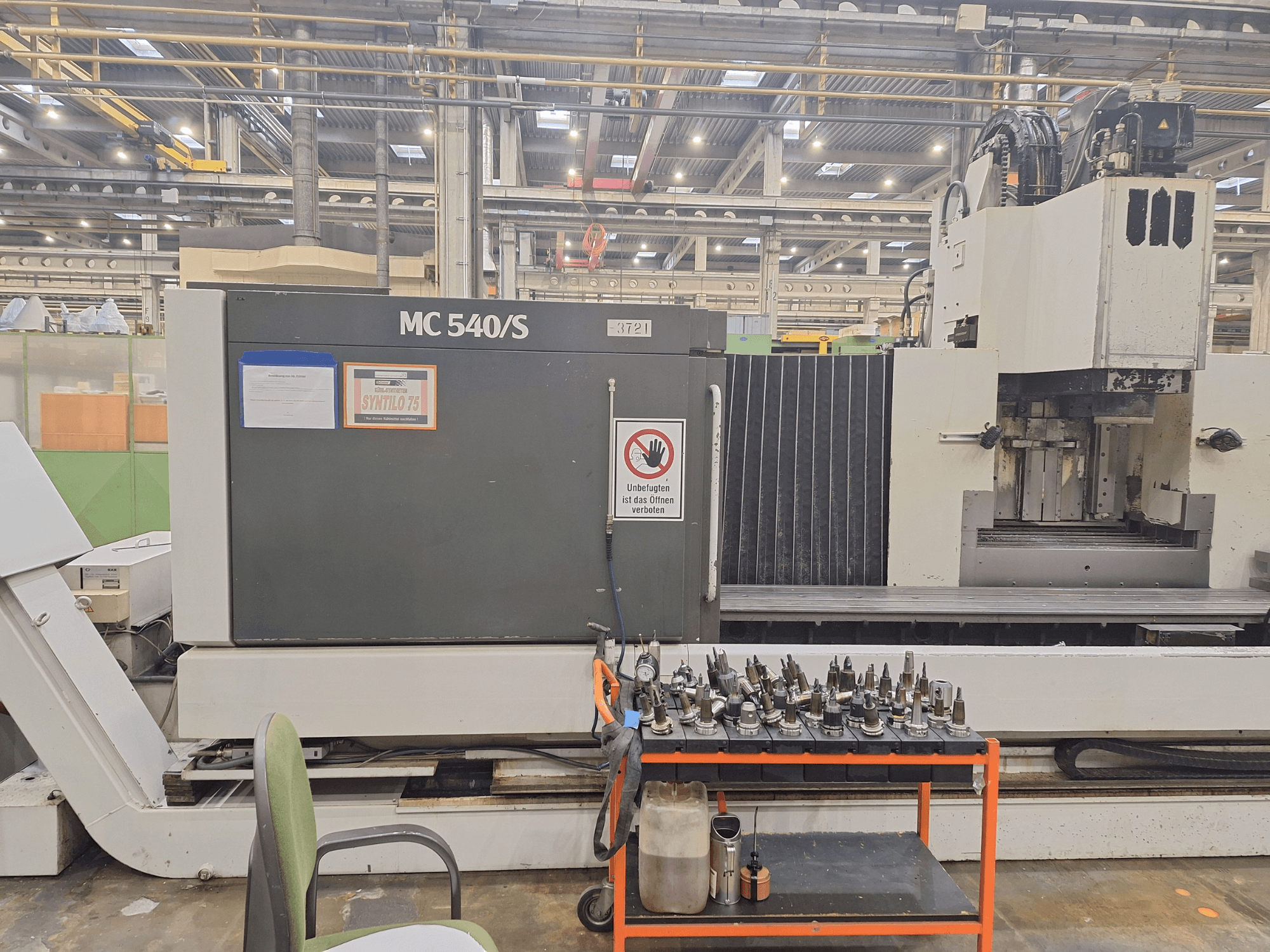 Vertikālais apstrādes centrs STAMA MC 540S darbnīcā, skats no sāniem ar instrumentu turētājiem uz ratiņiem blakus.