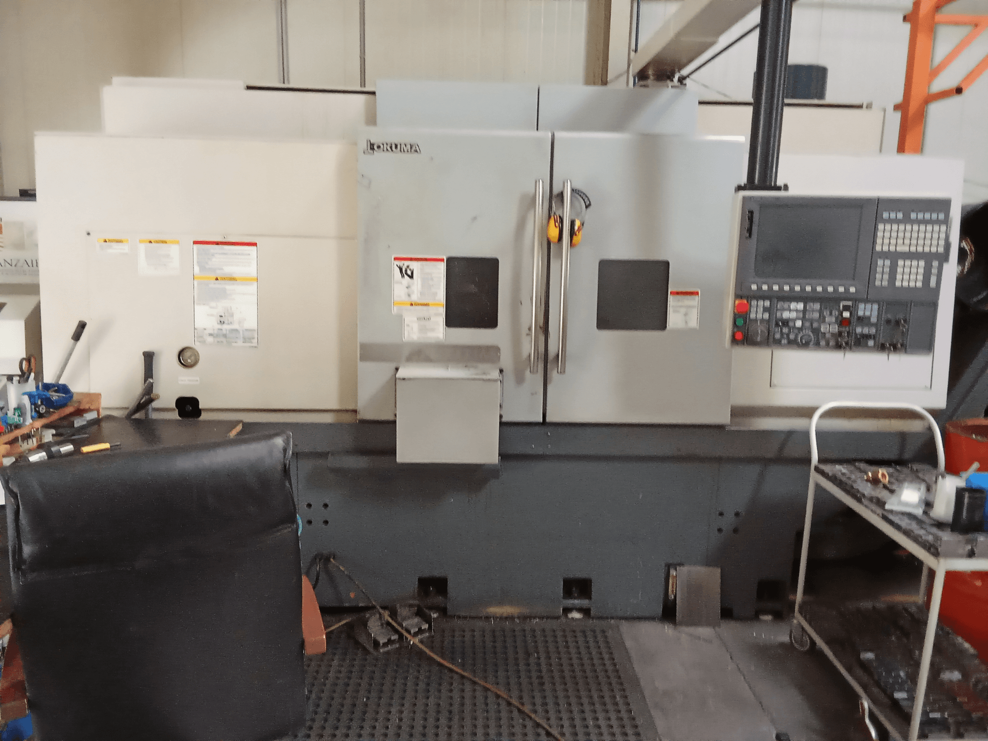 Okuma Genos L300 E-M horizontālās virpošanas iekārta priekšpusē, ar vadības paneli un dubultām durvīm, darbnīcas vidē.