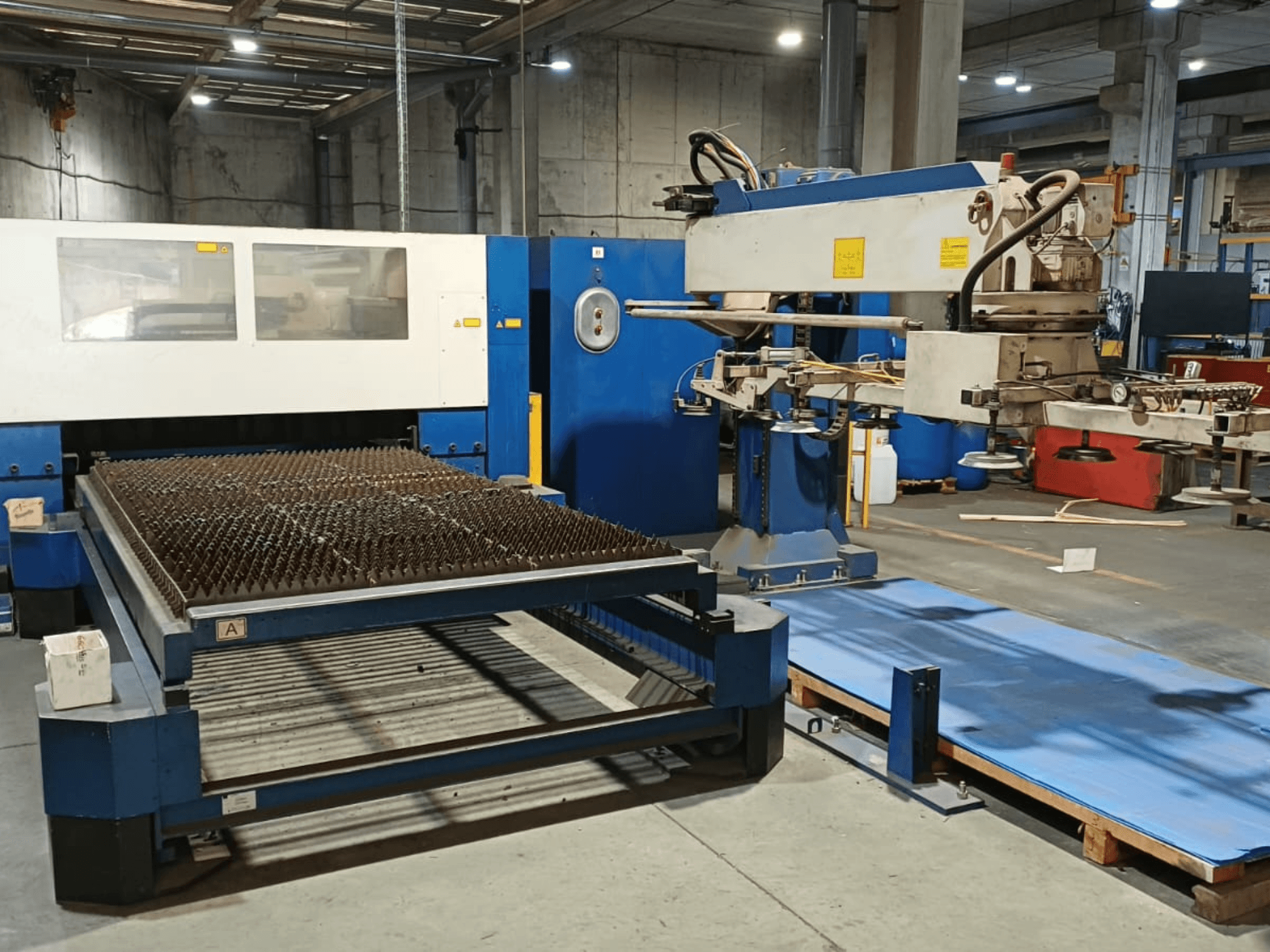 TRUMPF TRUMATIC L 3030 CO₂ lāzergriešanas iekārta, skats no sāniem, kurā redzams griešanas galds ar smailiem izciļņiem un robotizētā roka.