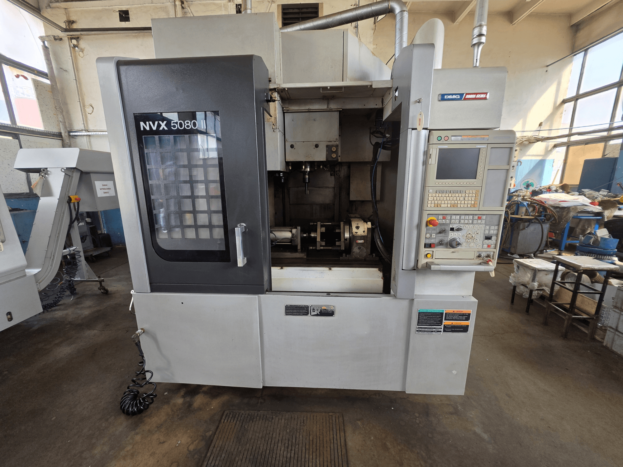 DMG Mori Seiki NVX 5080 II/40 vertikālā apstrādes centra skats no priekšpuses ar vadības paneli un caurspīdīgu logu, kas atklāj apstrādes zonu.