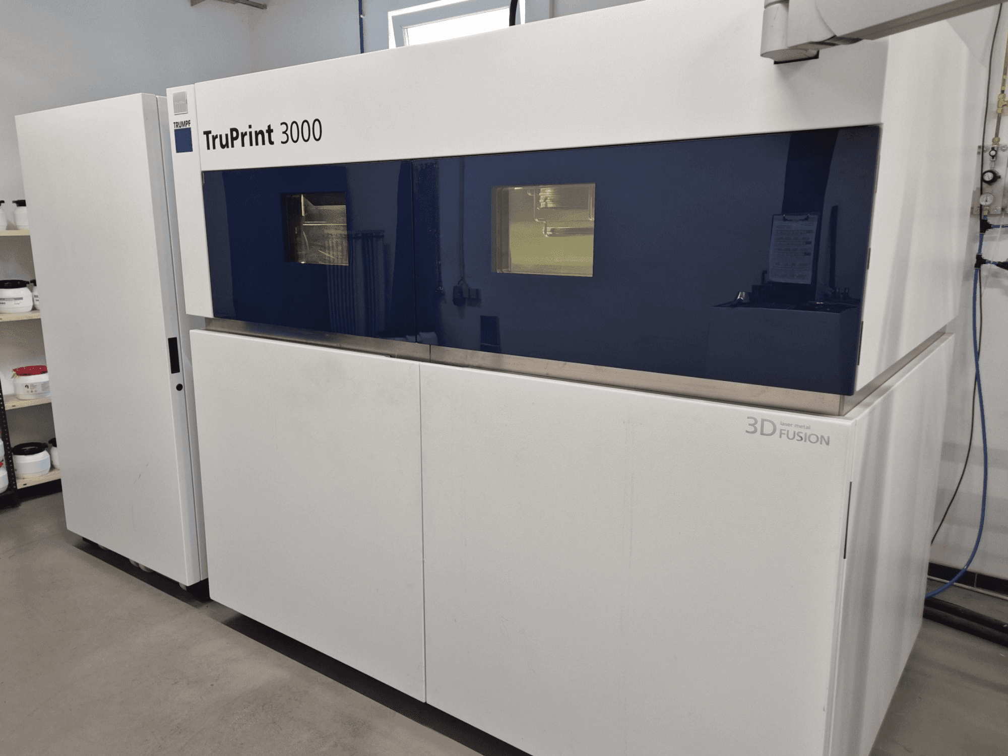 TruPrint 3000 metāla 3D printeris priekšpusē, ar zilu paneļu dizainu un lāzerterapstrādes tehnoloģiju.