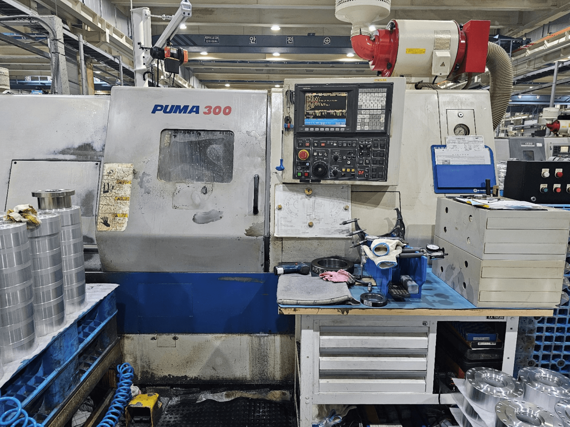 PUMA 300 CNC darbnīcā; priekšējais skats, kurā redzams vadības panelis, instrumenti un metāla detaļas uz darbgalda.