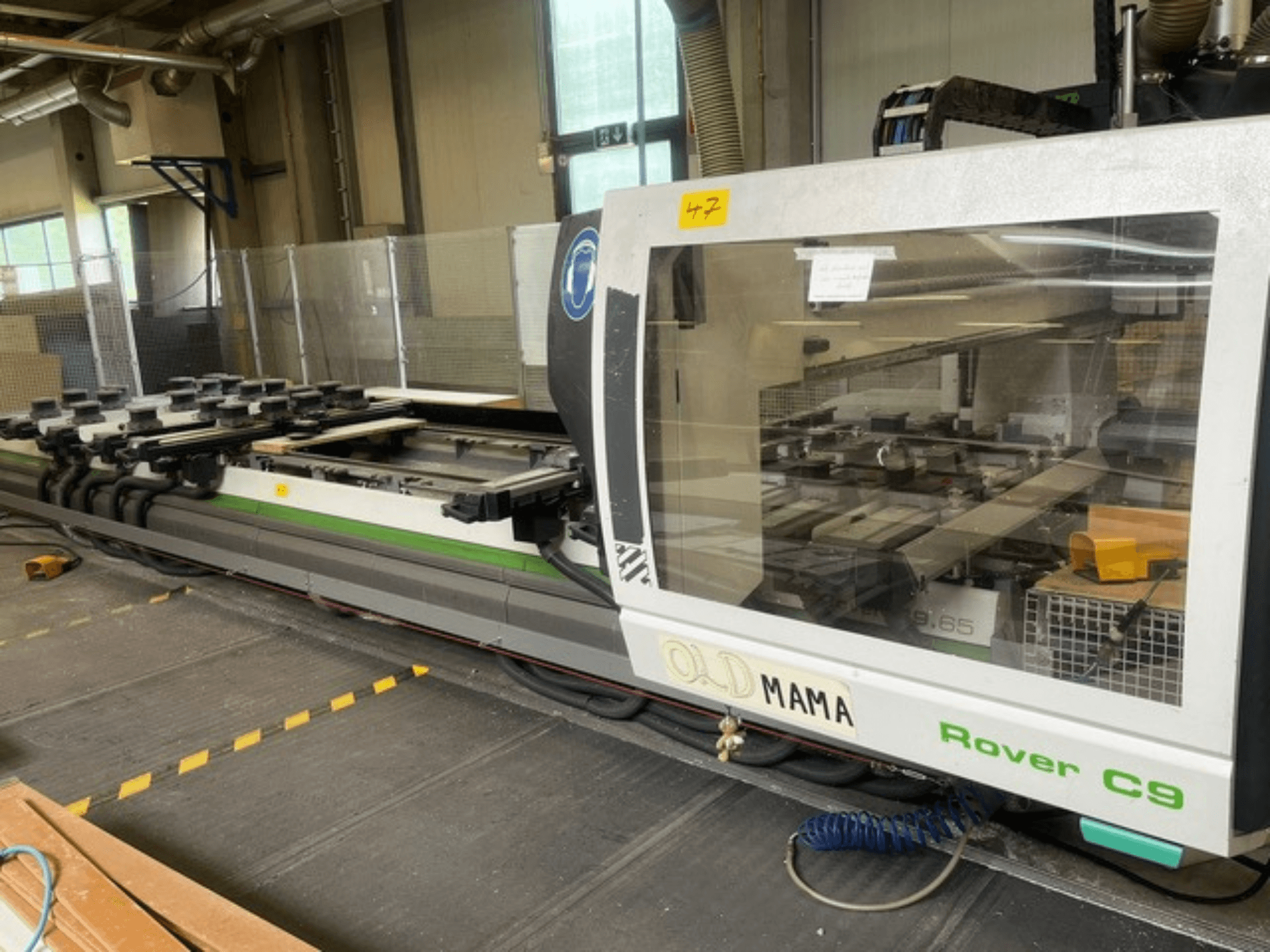 BIESSE Rover C 9.65 CNC frēzētājs darbnīcā, sānu skats, kurā redzams vadības panelis un darba galds ar skavām.
