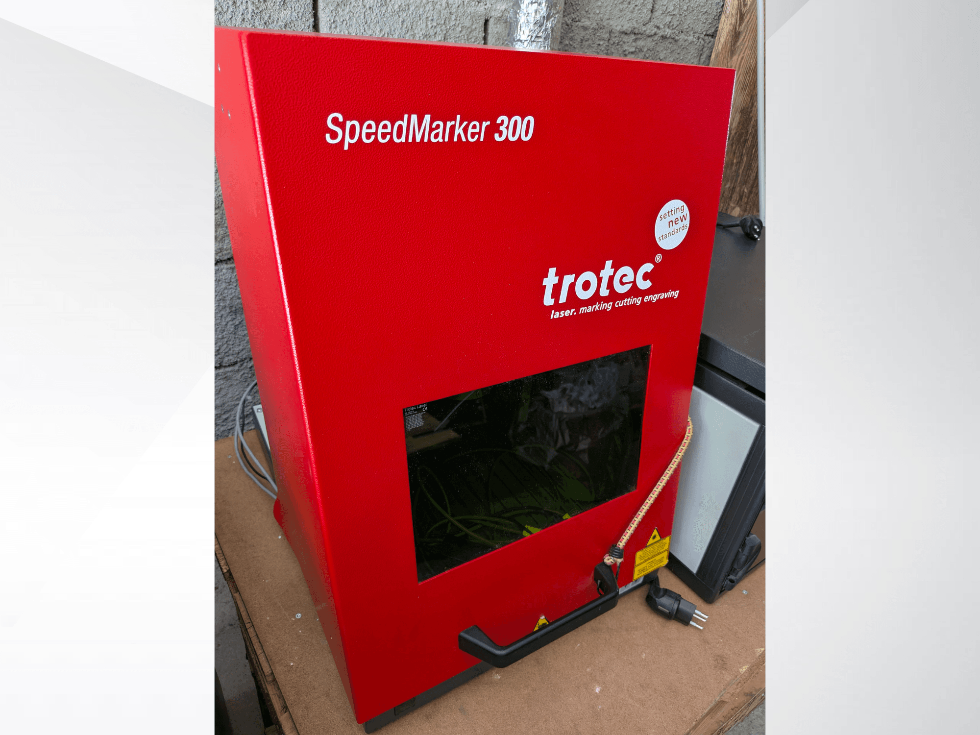 Sarkanā Trotec SpeedMarker 300 lāzera marķēšanas iekārta priekšpusē, ar lielu skatu logu un strāvas pieslēgumu.