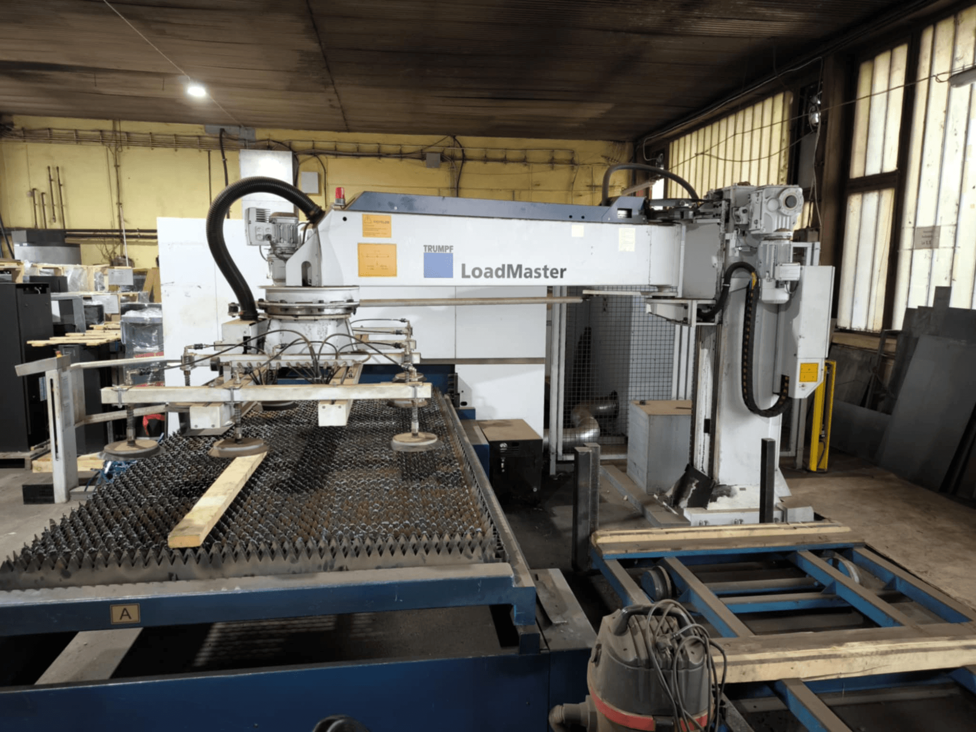 TRUMPF TRULASER 3030 FIBER griešanas iekārta, skats no priekšpuses, ar LoadMaster sistēmu un novietota uz režģa darba galda.