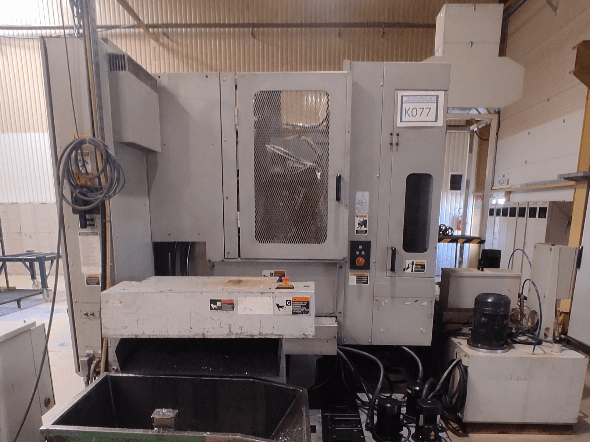 Pelēkas rūpnieciskās CNC darbgaldi ar lielu darba galdu, skats no sāniem, ar vadības paneli un dzesēšanas šķidruma rezervuāru, modelis K077.