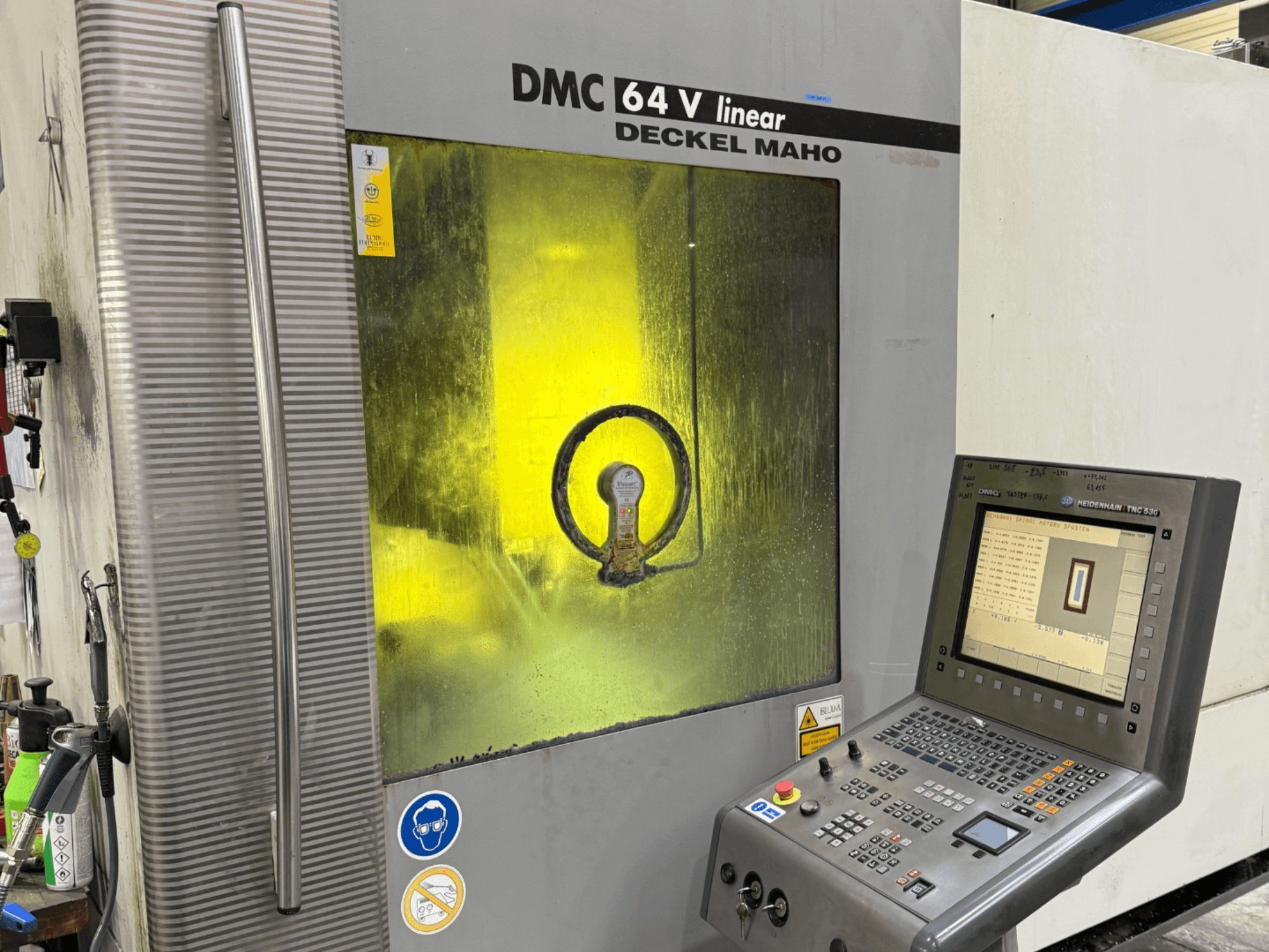 Deckel Maho DMC 64 V lineārā, priekšējais skats uz mašīnu ar vadības paneli un dzelteni tonētu logu, kurā redzami iekšējie mehānismi.