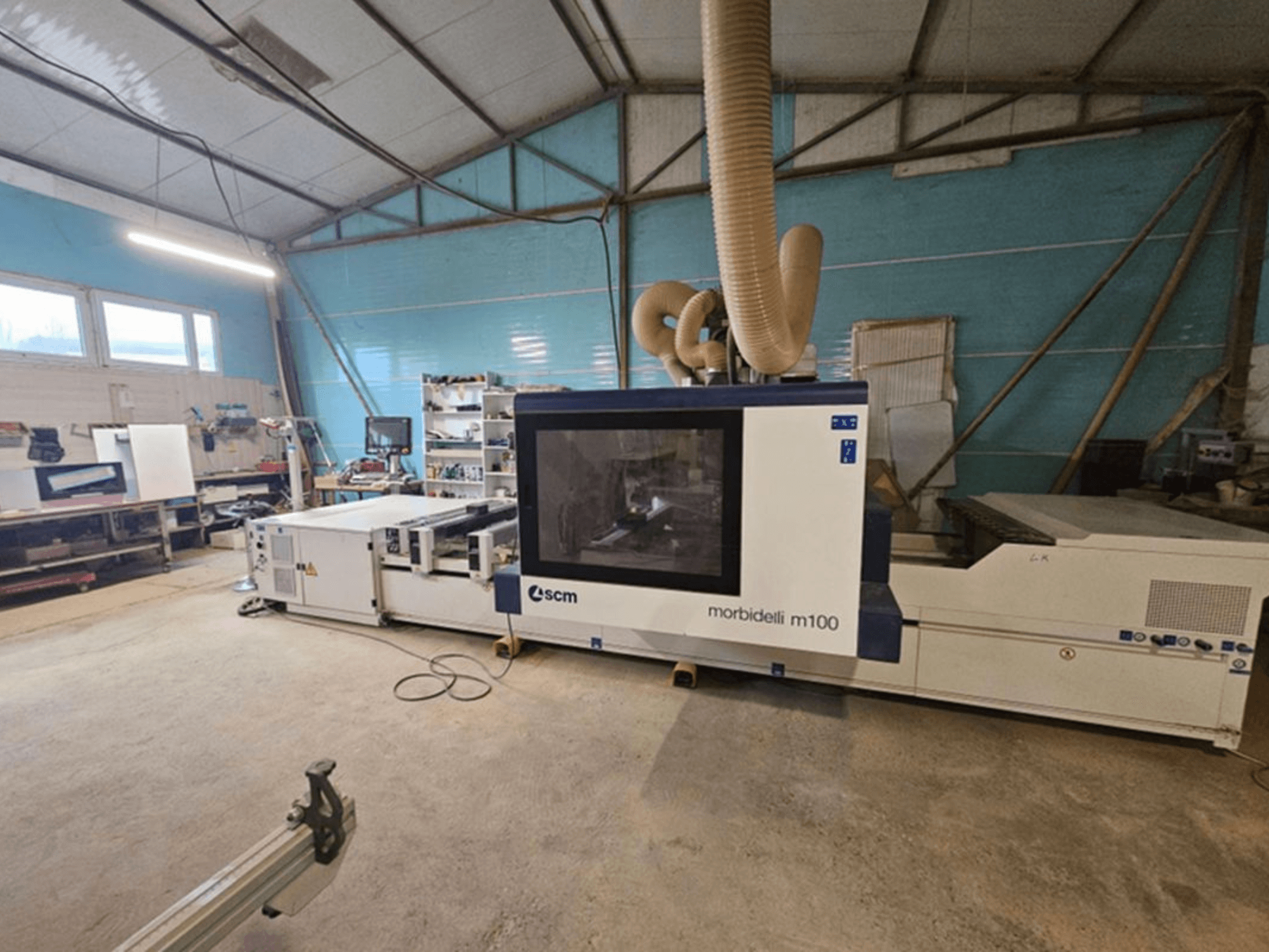 SCM MORBIDELLI M 100 CNC apstrādes centrs darbnīcā, skats no priekšpuses ar putekļu nosūces sistēmu un darba vietas aprīkojumu.