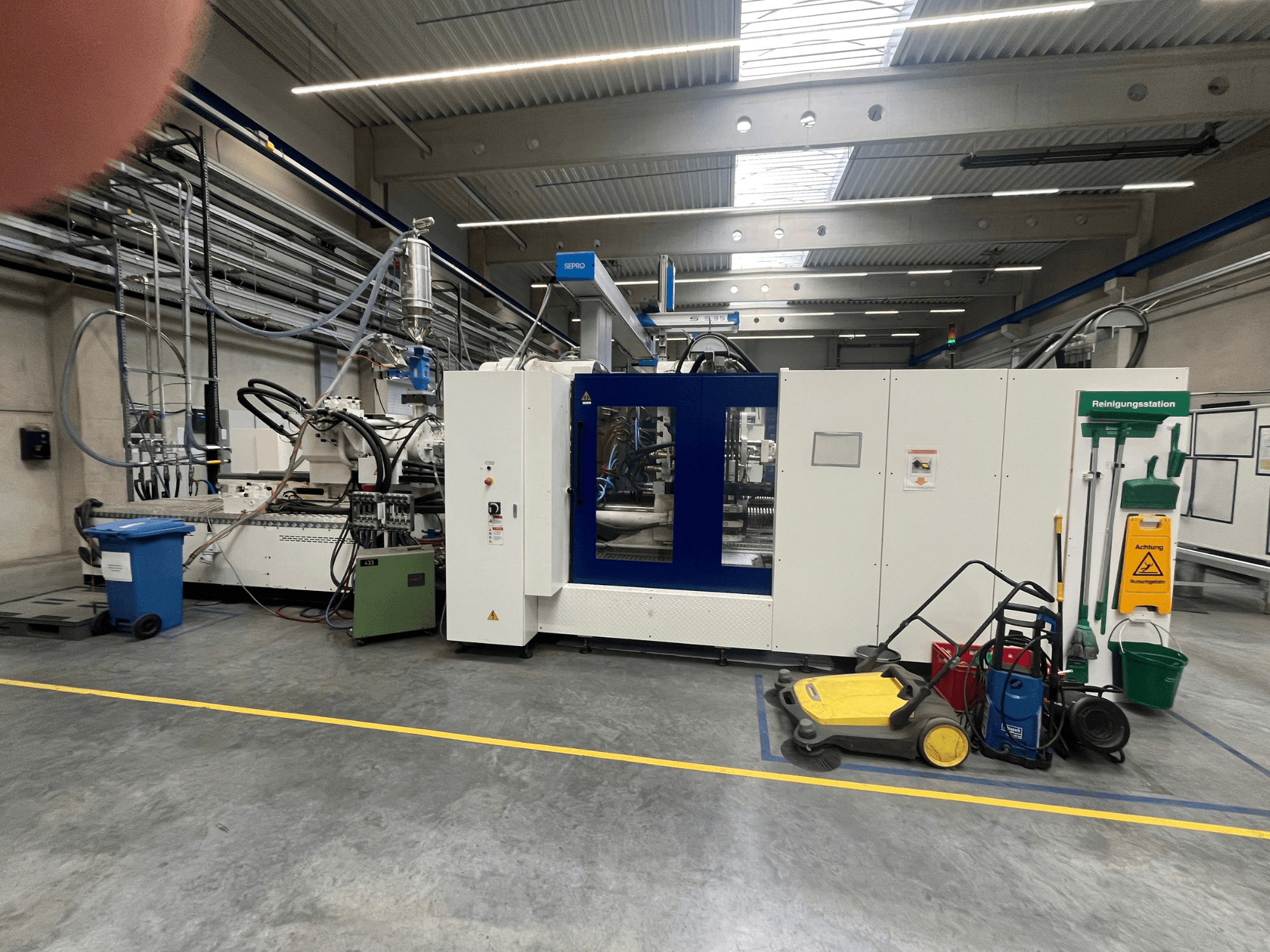 Rūpnieciska iekārta rūpnīcā, skats no priekšpuses, ar ziliem un baltiem paneļiem, šļūtenēm un tīrīšanas iekārtām blakus.