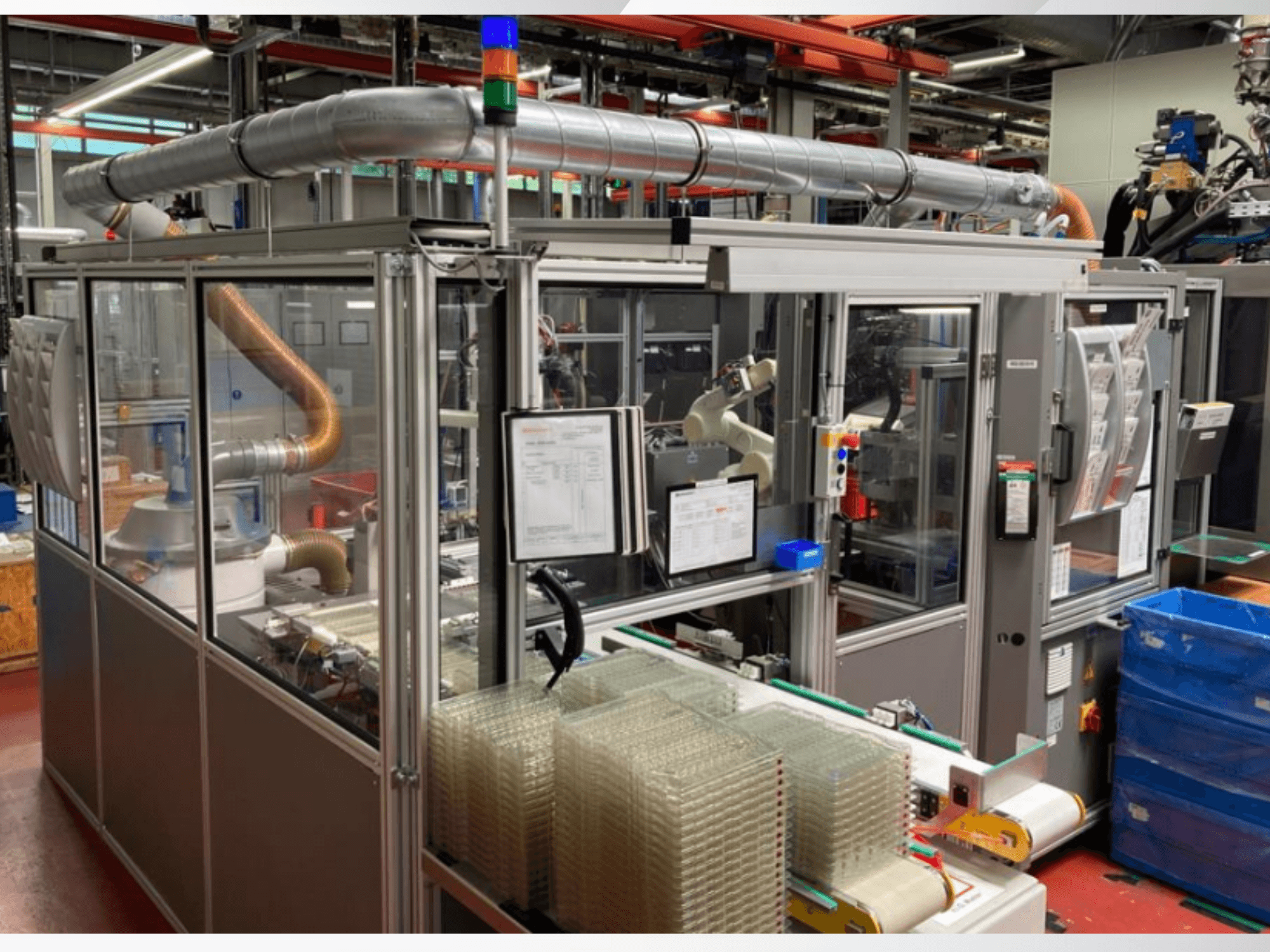 Skats no priekšpuses uz automatizētu iekārtu ar metāla caurulēm, vadības paneli un sakrautiem caurspīdīgiem plastmasas traukiem uz rūpnīcas grīdas.