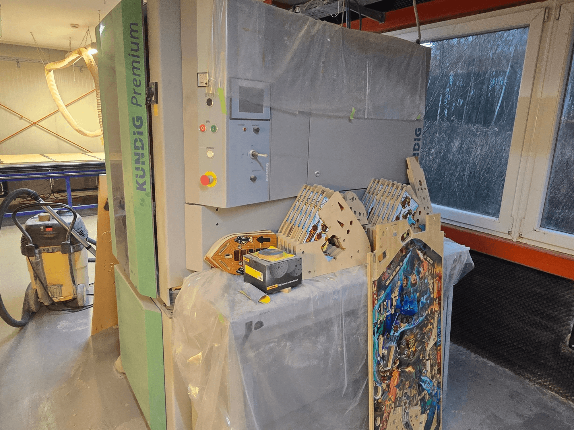 Kundig Premium-1 1100-C lentes slīpmašīna darbnīcā, redzams vadības panelis un koka daļas, skats no sāniem, fonā putekļu putekļu putekļu sūcējs.