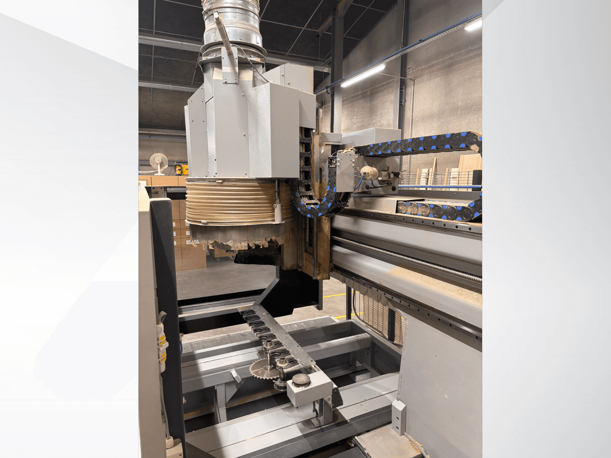 CNC frēzmašīna ar lielu vārpstu un konveijeru, skats no malas, kurā redzami kokapstrādes instrumenti un iekārtu sastāvdaļas.